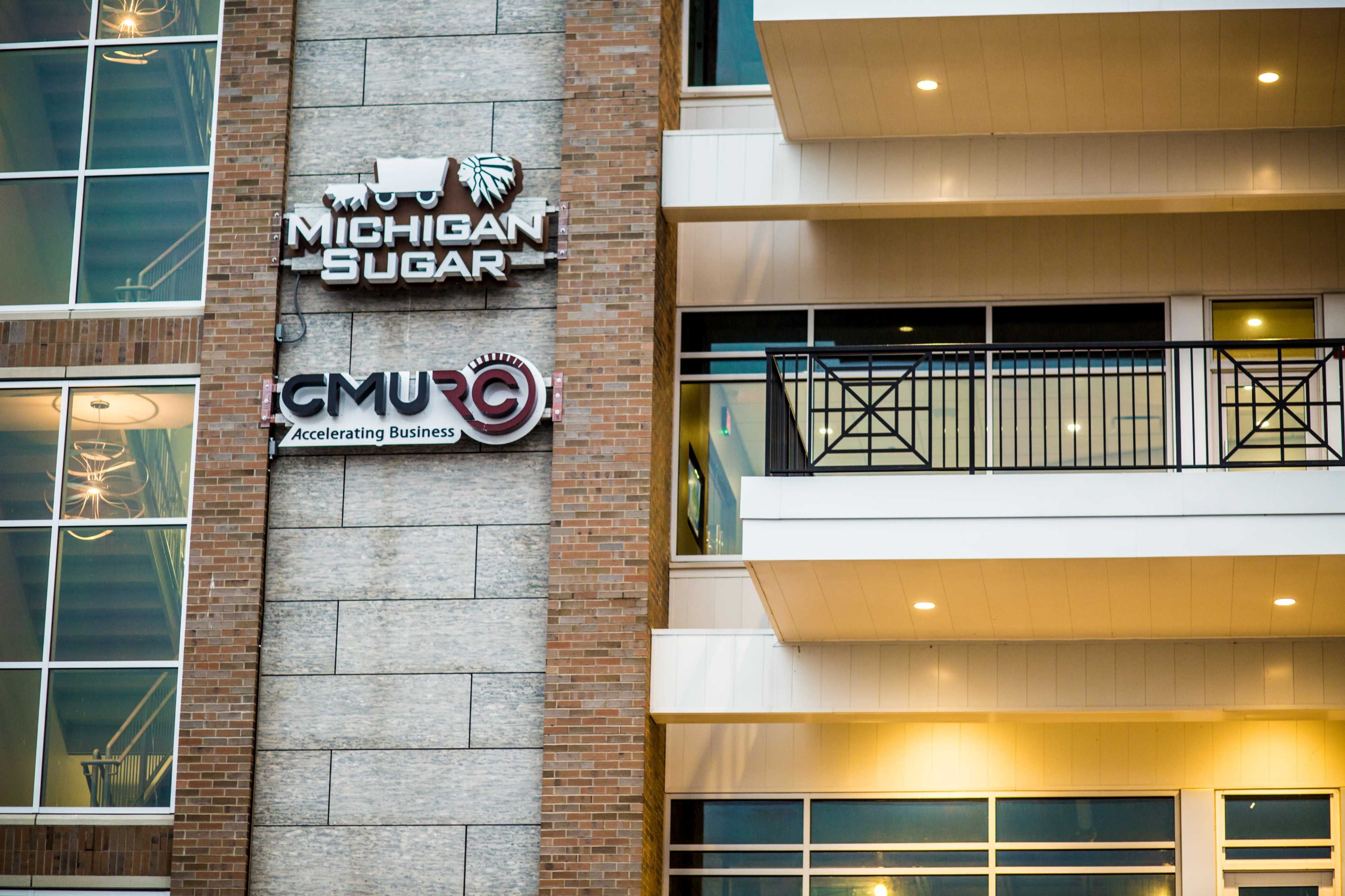 The image shows the exterior of a building featuring a sign for Michigan Sugar and another sign for CMU, along with a balcony on the upper floor.