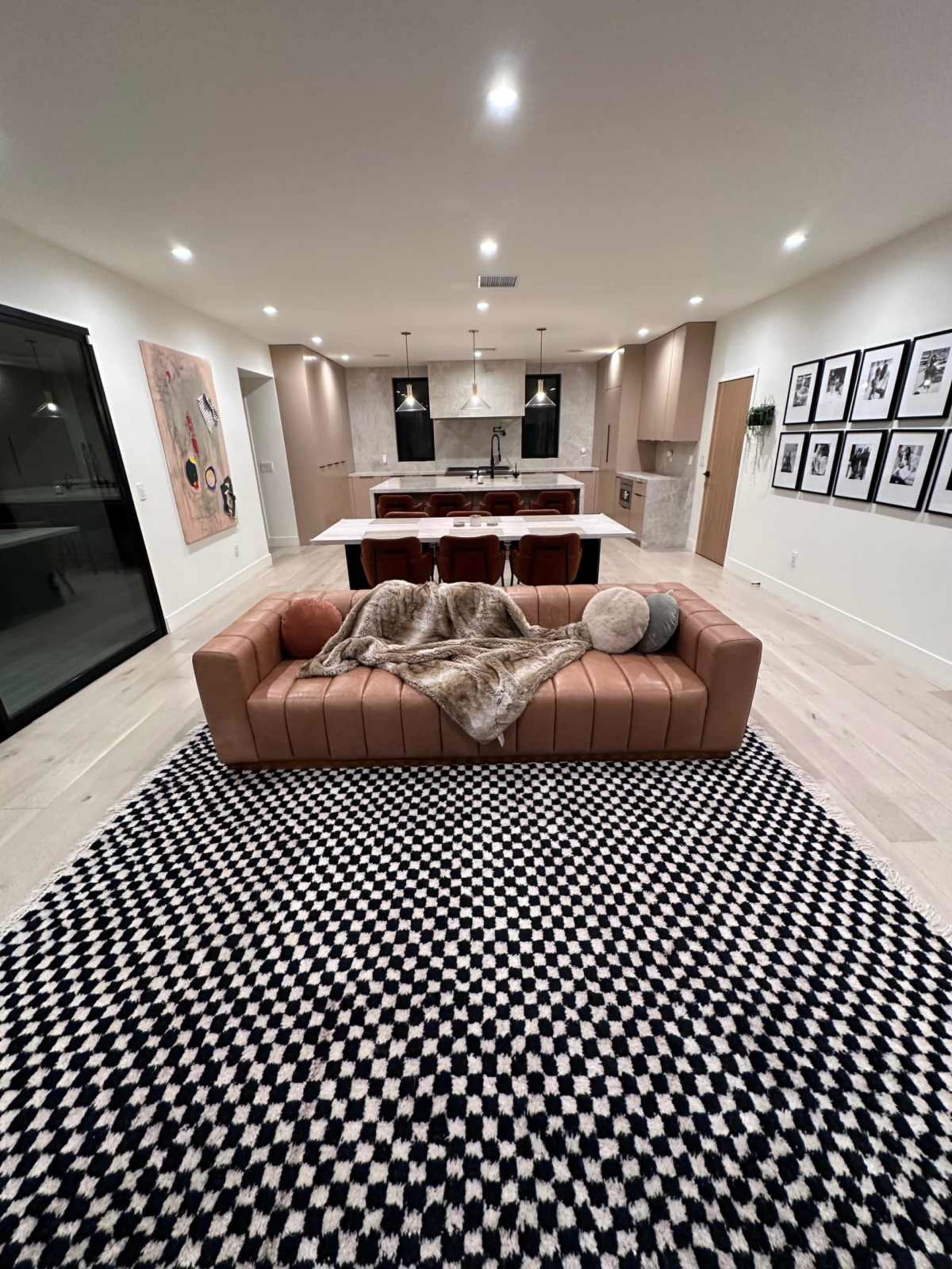 The image shows a modern living room with a tan leather sofa, a patterned black and white rug, and a kitchen area in the background featuring bar stools and artwork on the walls.