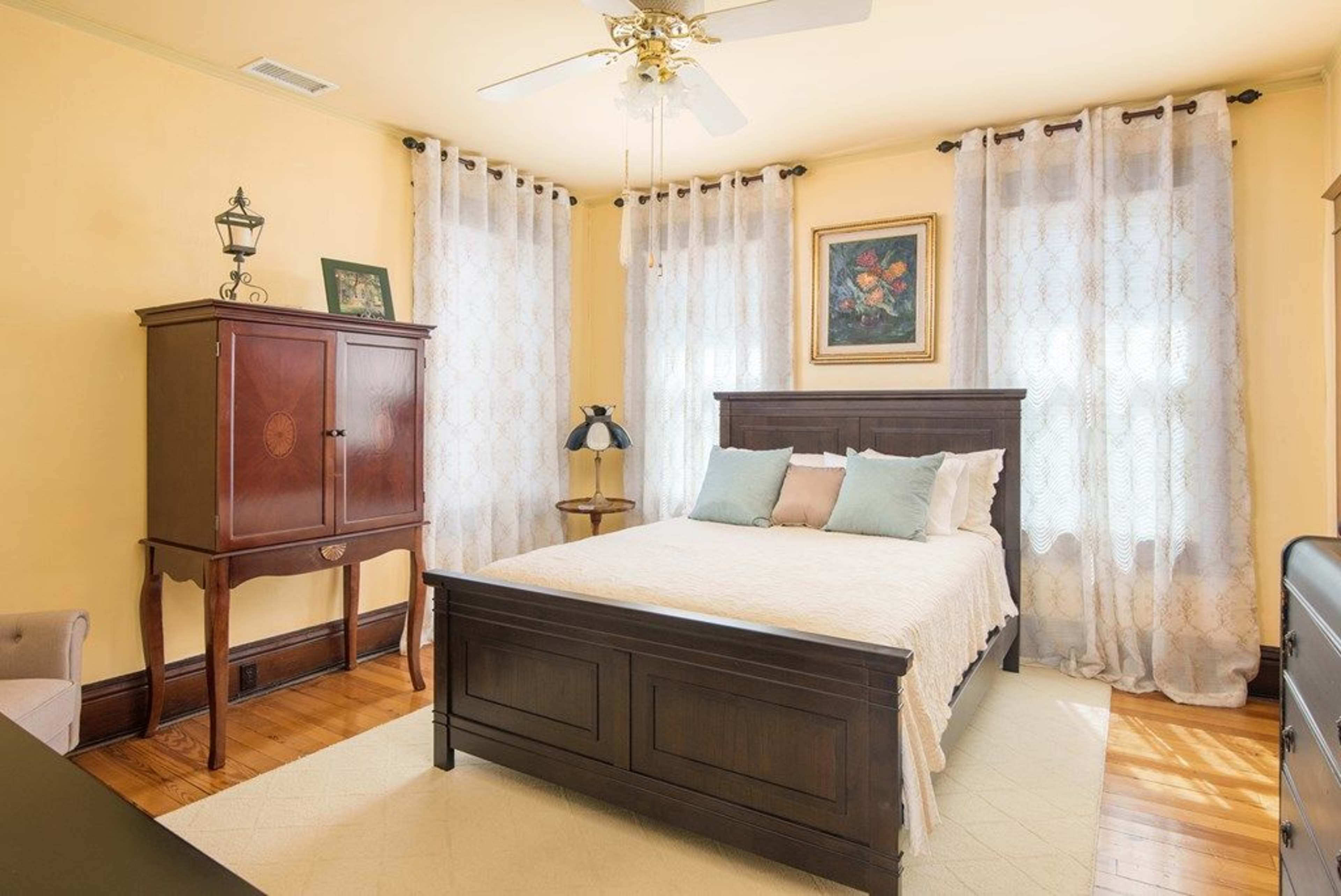 The image shows a well-furnished bedroom featuring a wooden bed, an armoire, a decorative lamp, and sheer curtains allowing light to filter in.