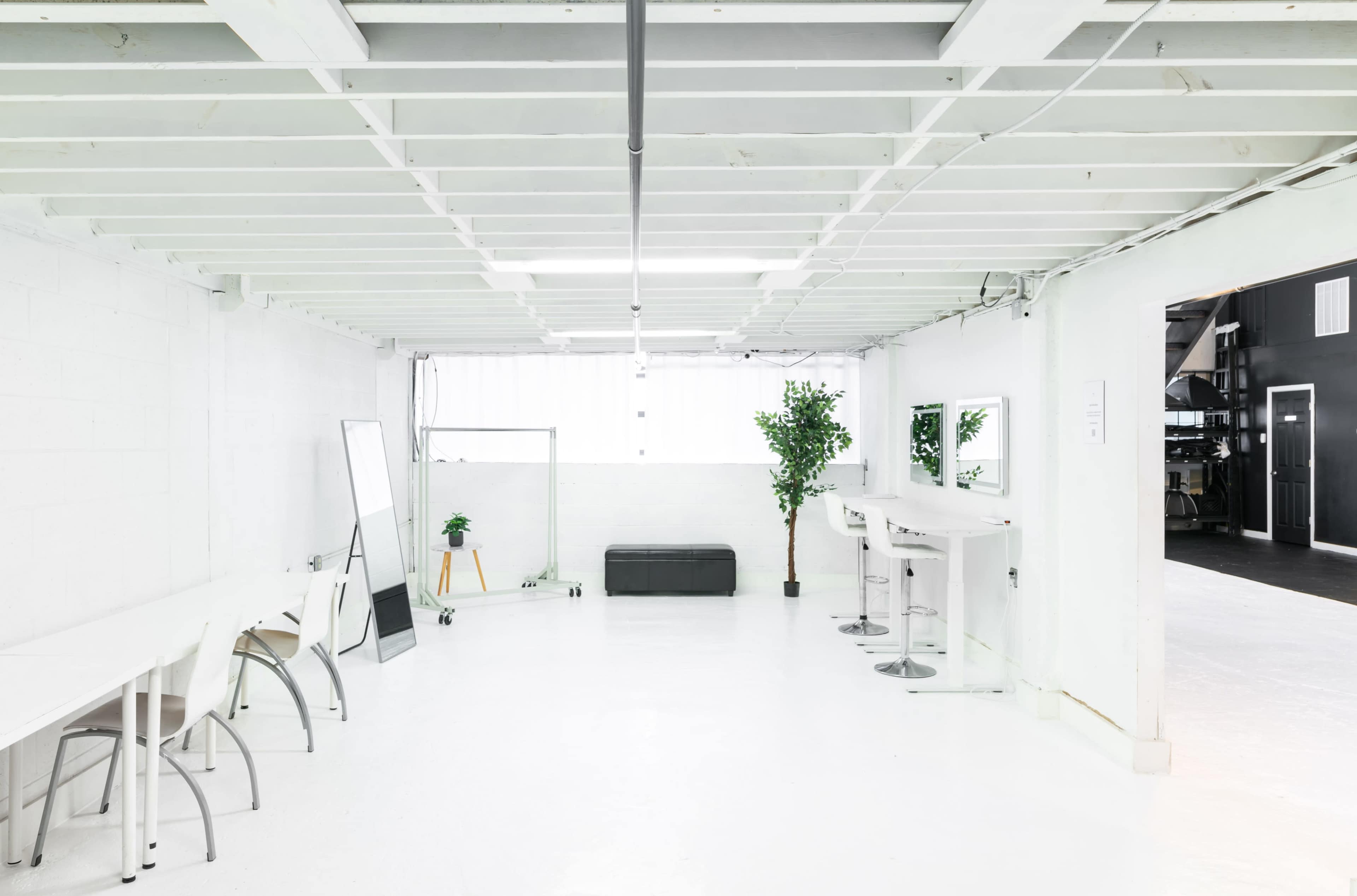 The image shows a spacious, well-lit white interior with minimalist furnishings, including a table, chairs, a mirror, and a plant.