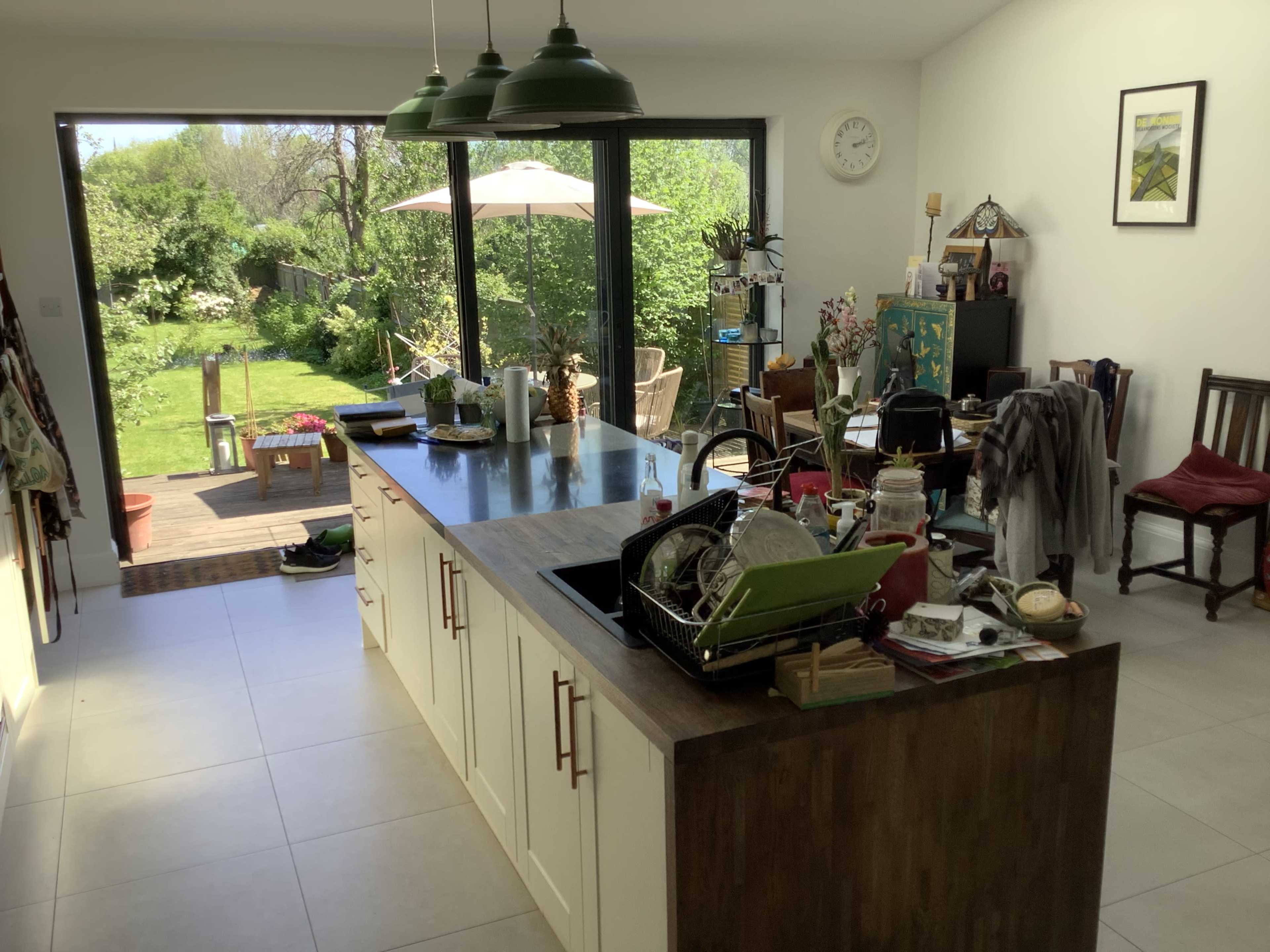 A spacious kitchen with a central island, cluttered countertops, and large glass doors leading to a garden.