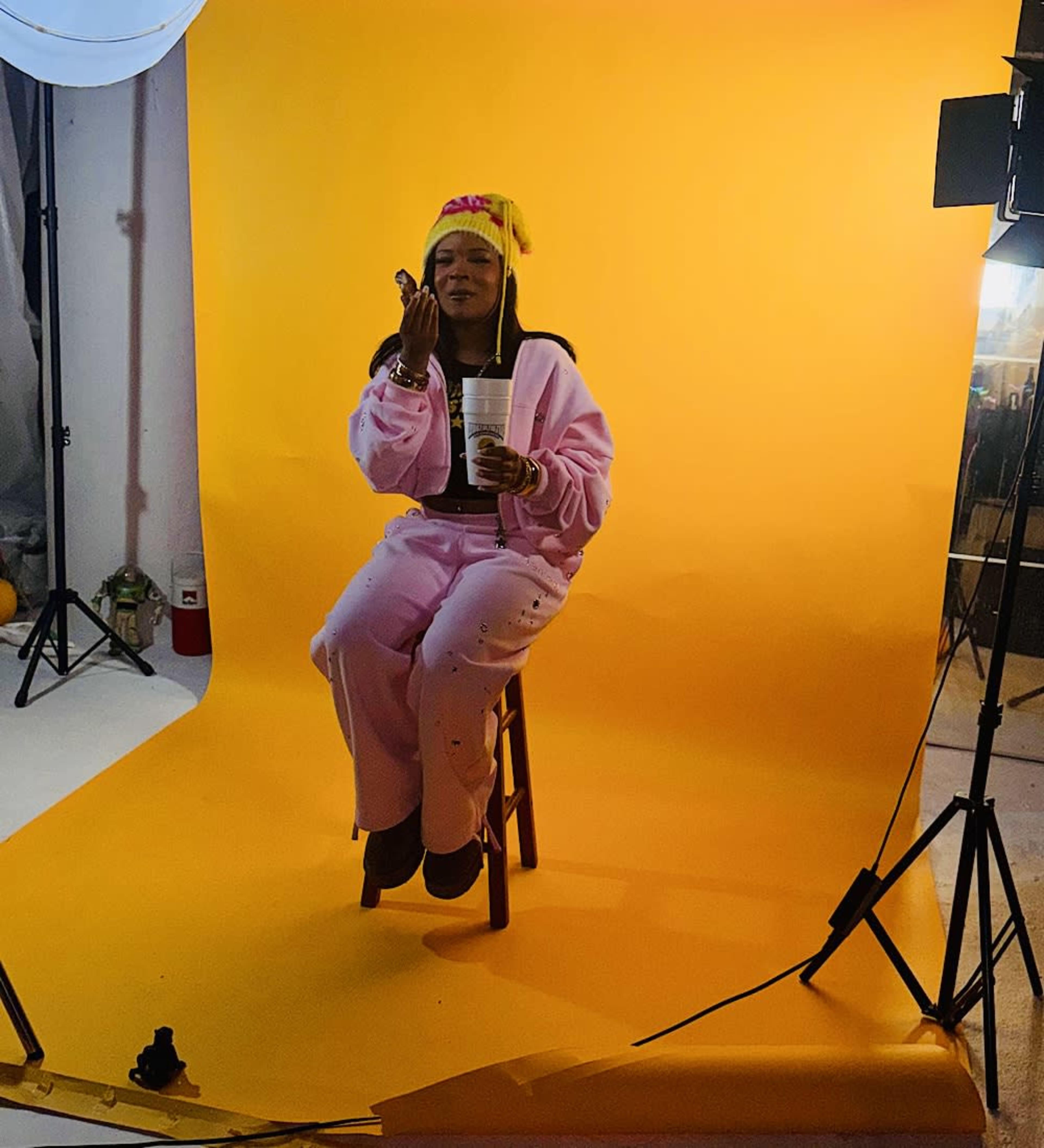 A person sitting on a stool in a pink outfit holds a drink against a bright yellow background.
