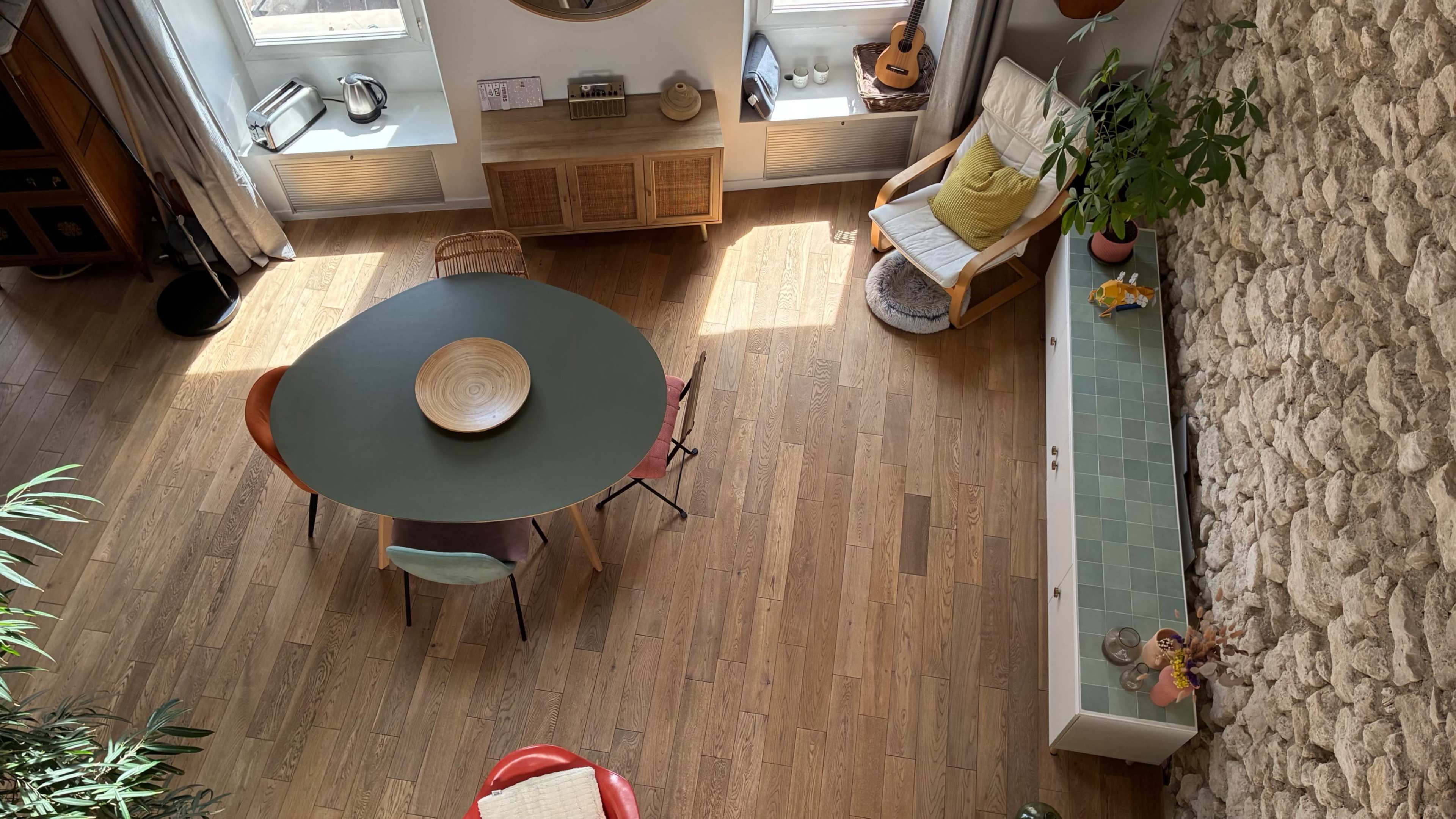 The image shows a spacious, minimalist living area featuring a round dining table with colorful chairs, natural wood flooring, and a stone accent wall, illuminated by natural light from large windows.