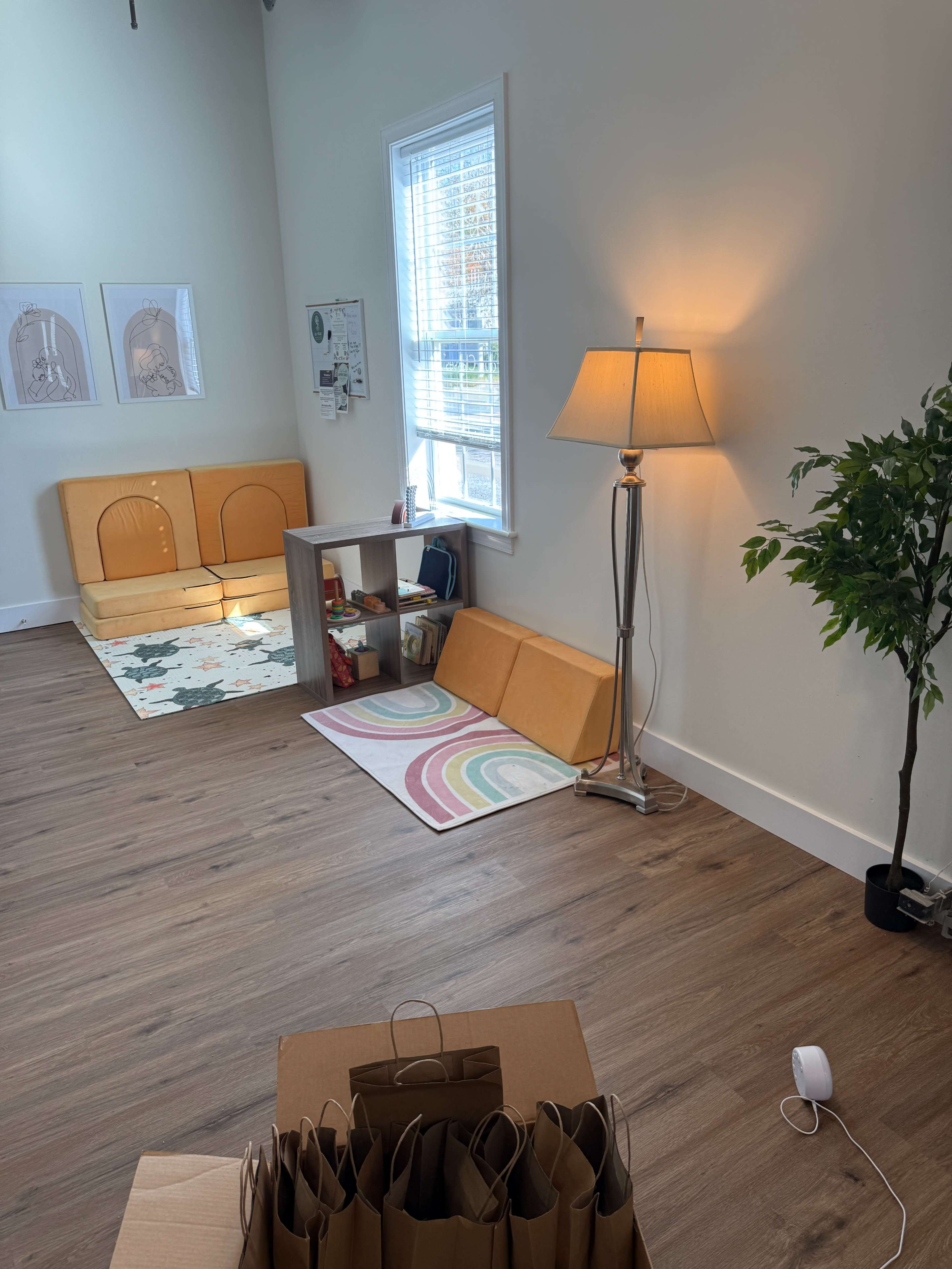 The image shows an interior room furnished with a lamp, a small table, and seating areas, along with a colorful rug and a stack of cardboard boxes on the floor.
