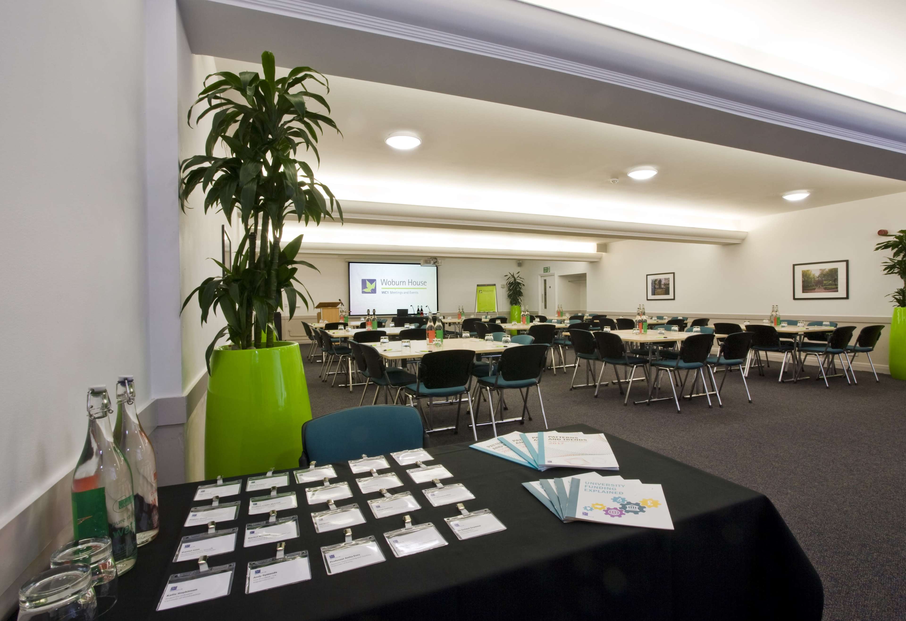 The image shows a conference room arranged with tables and chairs, featuring a registration table with name tags and a projector screen in the background.