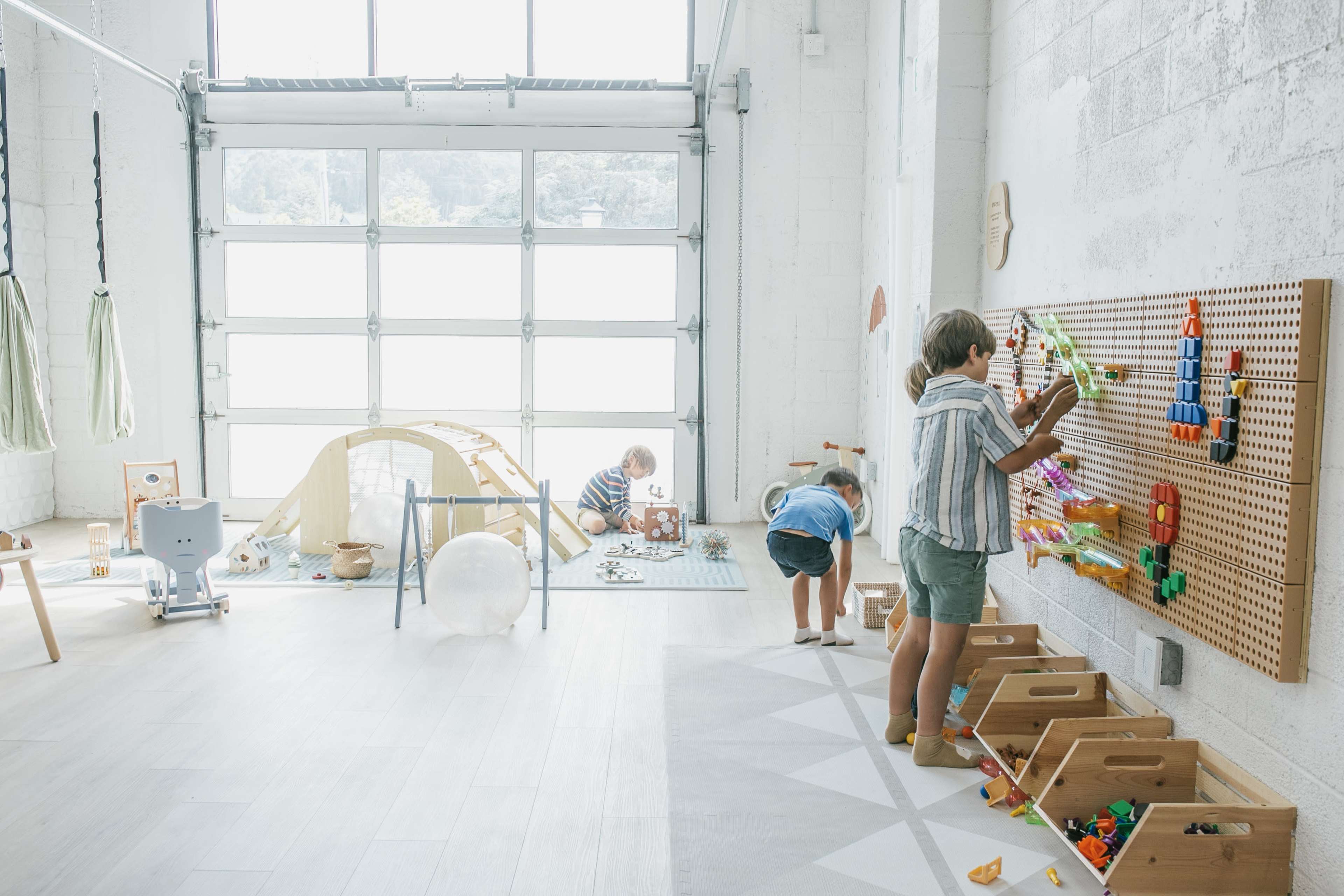 A brightly lit playroom features three children engaged in different activities, with one building on a pegboard, another playing with toys on the floor, and a fort made of fabric in the background.
