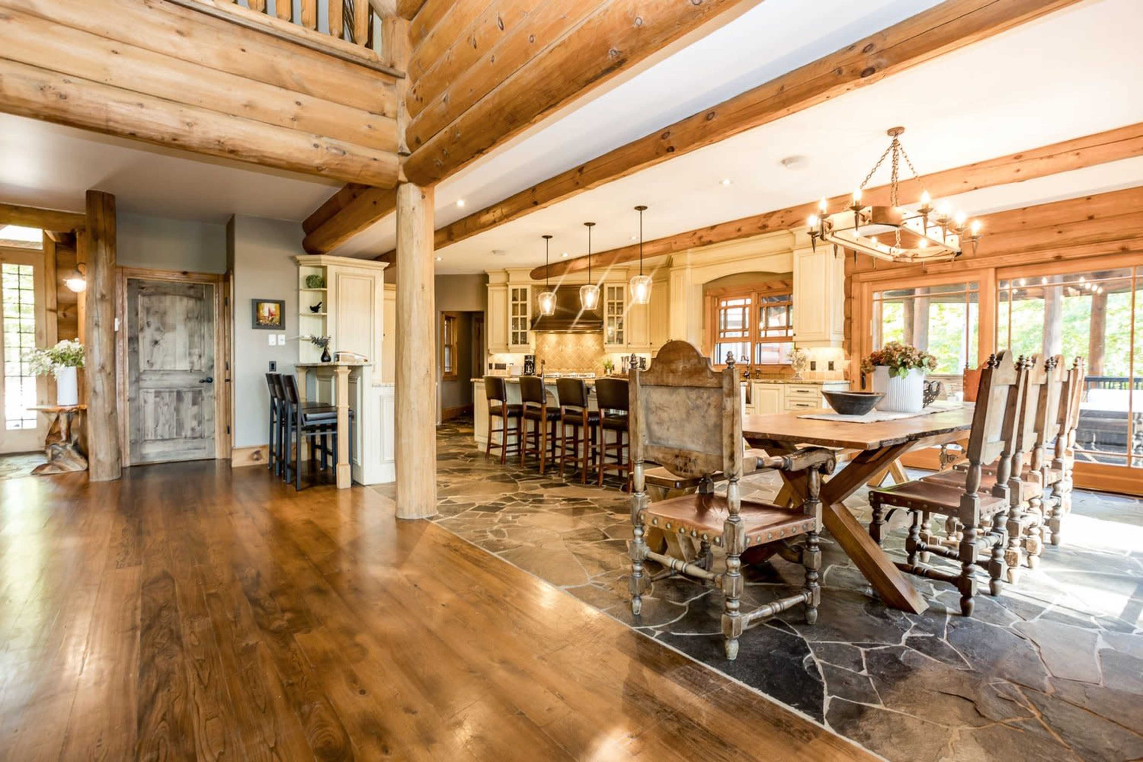 The image depicts a spacious, rustic dining area featuring a large wooden table surrounded by chairs, with a modern kitchen visible in the background.