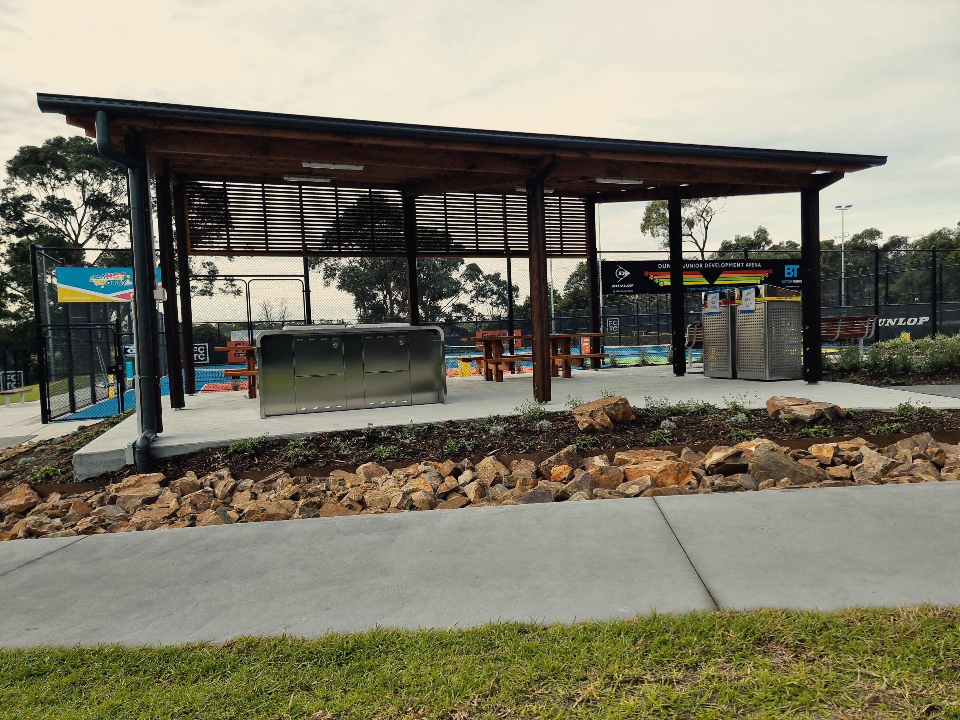 A covered outdoor area with a picnic table, a barbecue grill, and a landscaped stone border near a sports facility.