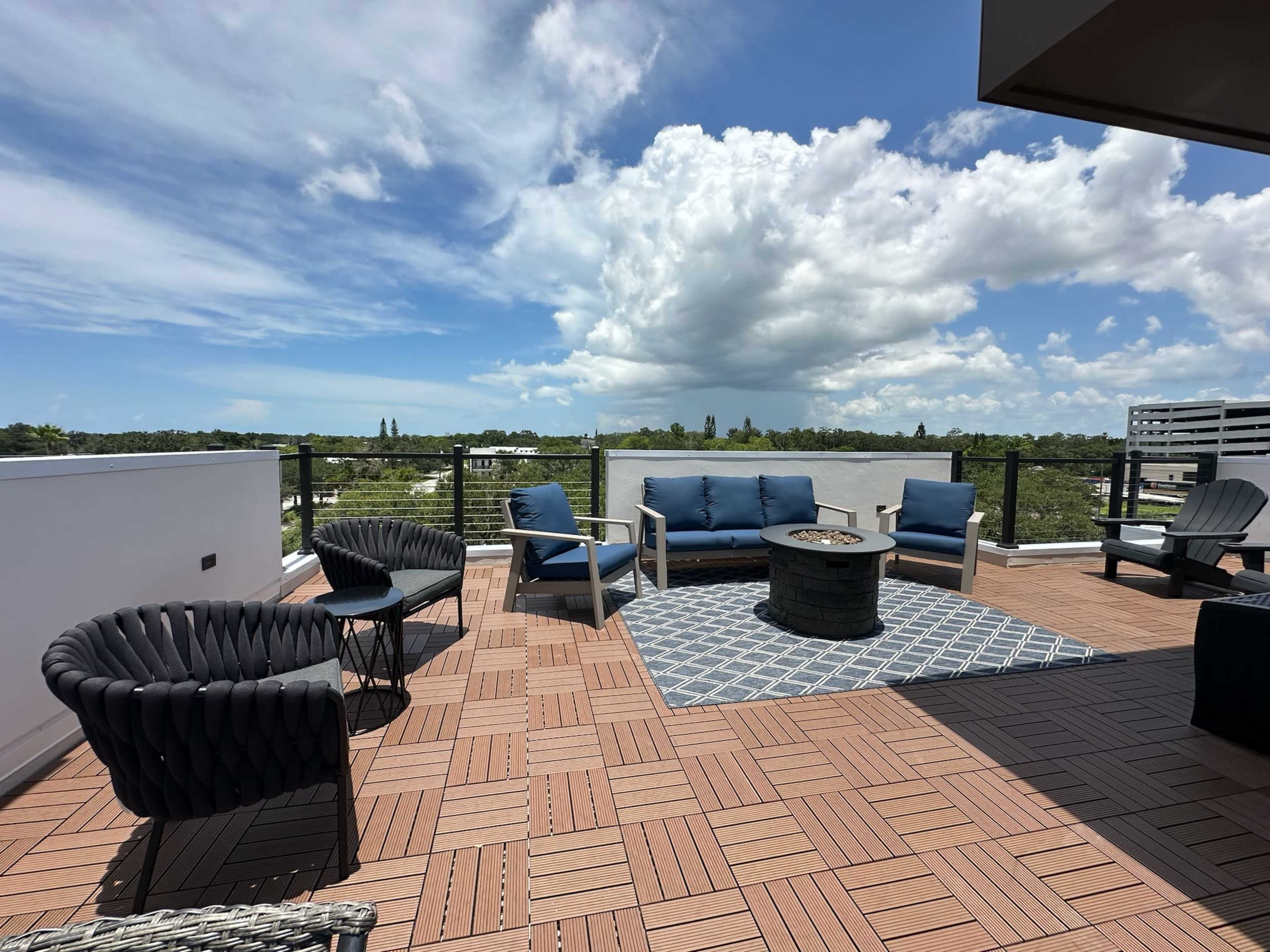 A rooftop terrace with outdoor seating, a round fire pit, and a clear blue sky with clouds overhead.