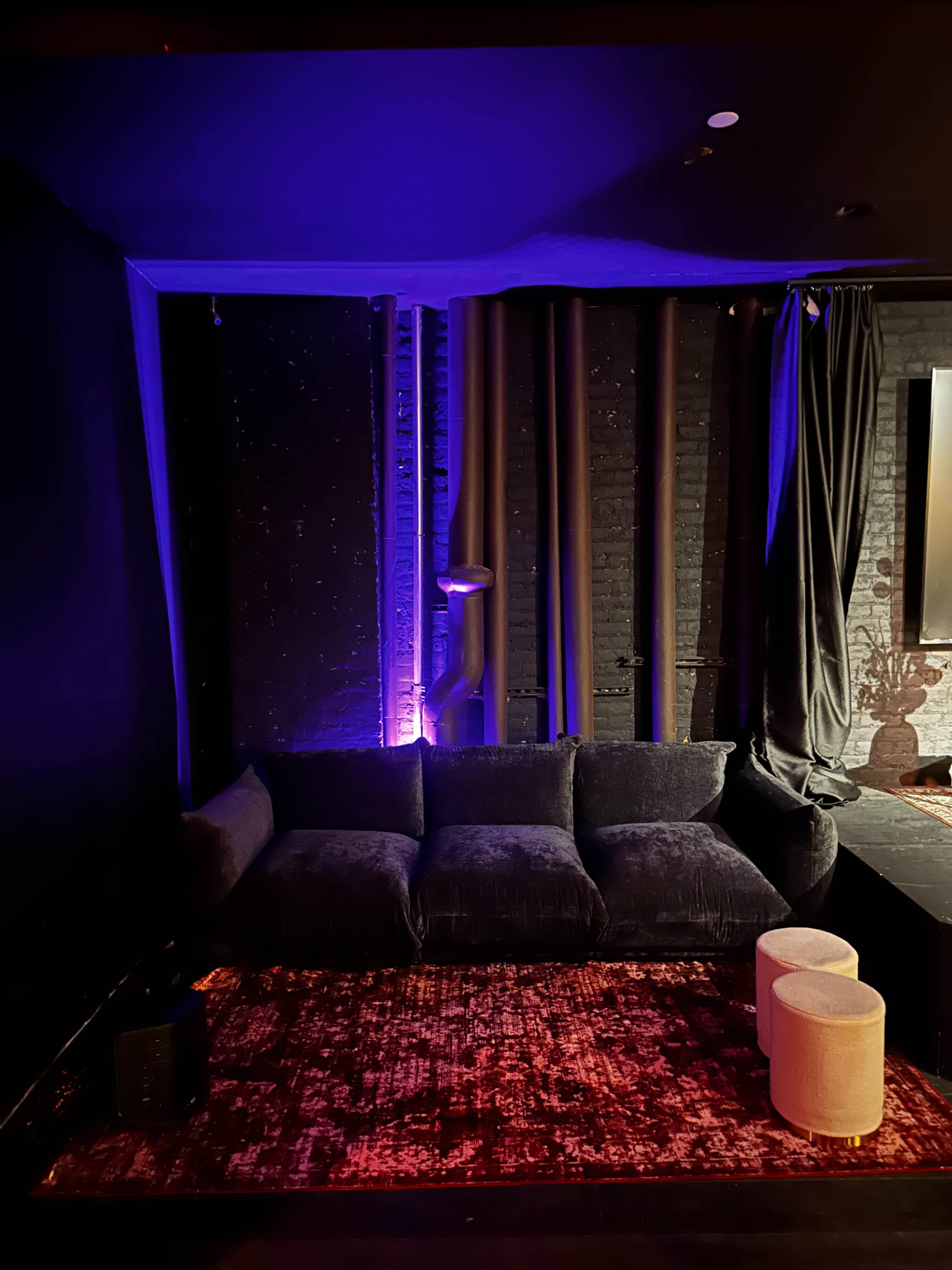 The image shows a dark room featuring a plush black sofa, a small round table, and purple ambient lighting against a brick wall.