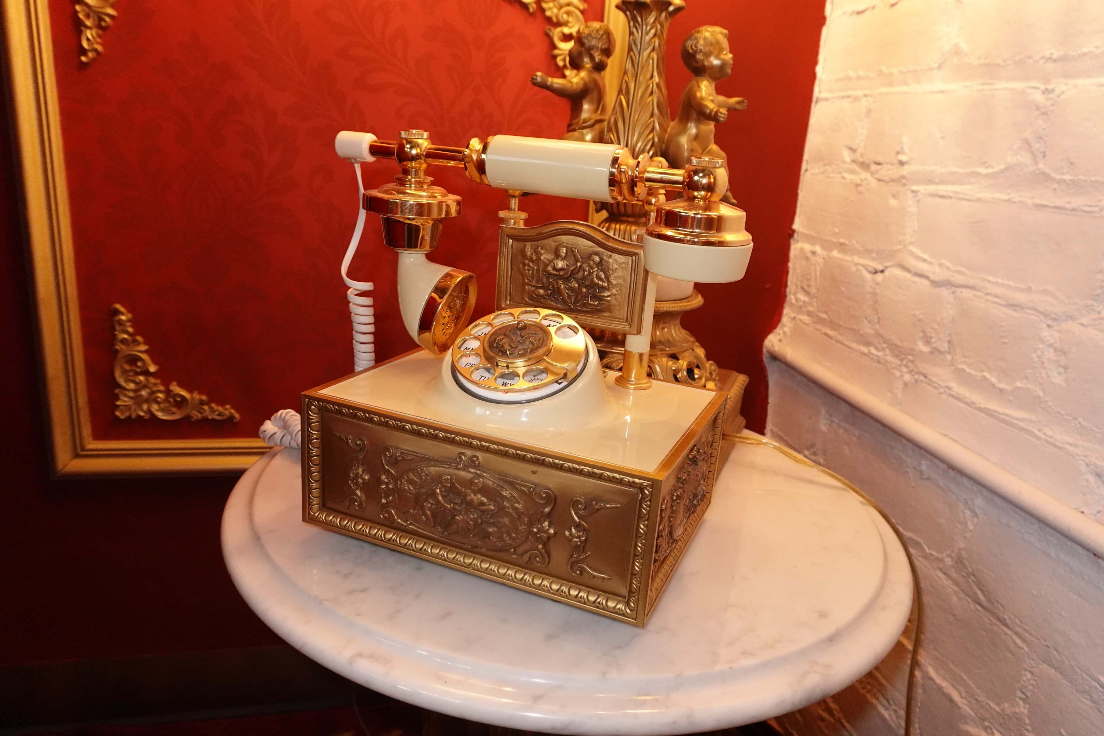 The image shows a vintage rotary telephone with a decorative design, placed on a marble-topped table against a red wall with ornate decor.