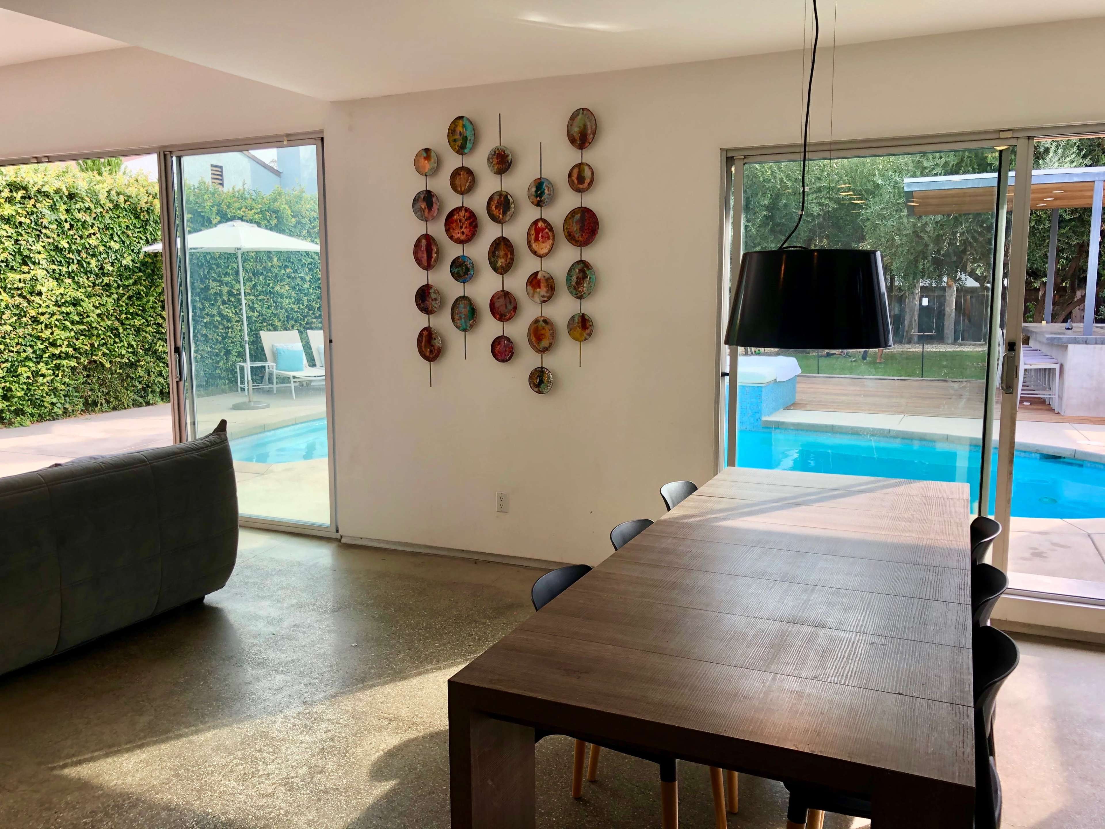 The image shows a modern dining area with a wooden table, black chairs, and a wall decorated with colorful plates, overlooking a swimming pool through large windows.