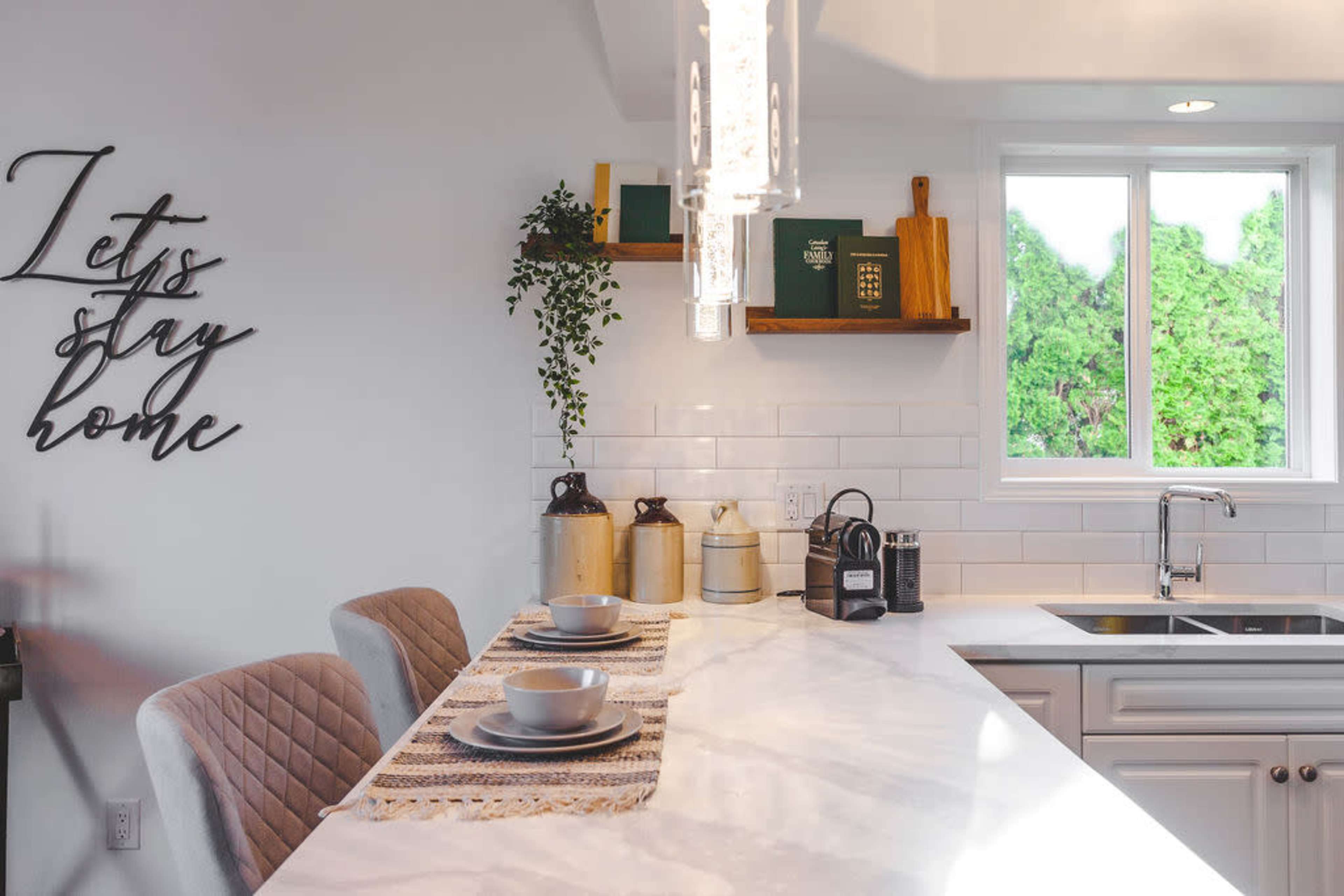 A modern kitchen features a marble countertop with two place settings, decorative canisters, and a window overlooking greenery.
