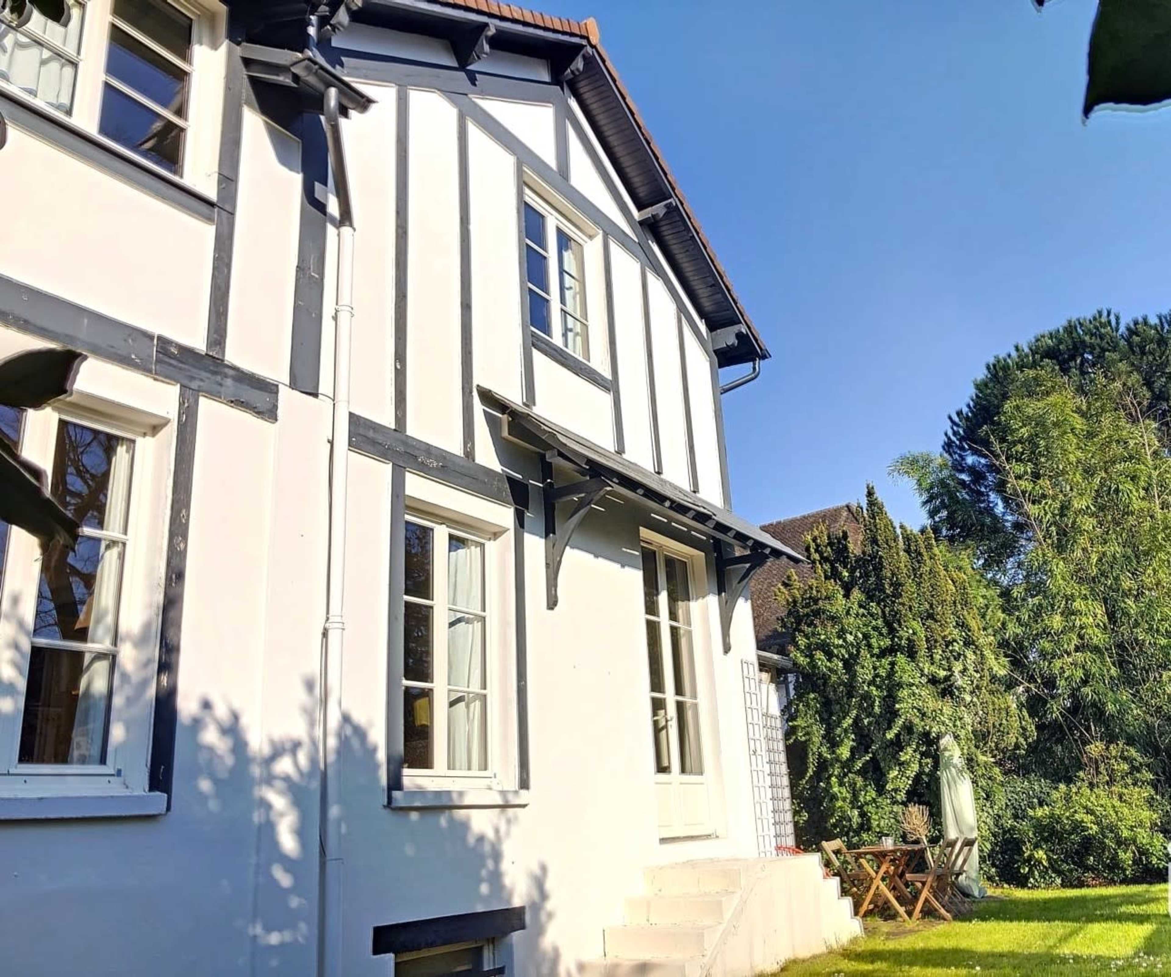 The house features a traditional timber-frame design with white walls and a manicured lawn, accompanied by a small seating area under a tree.