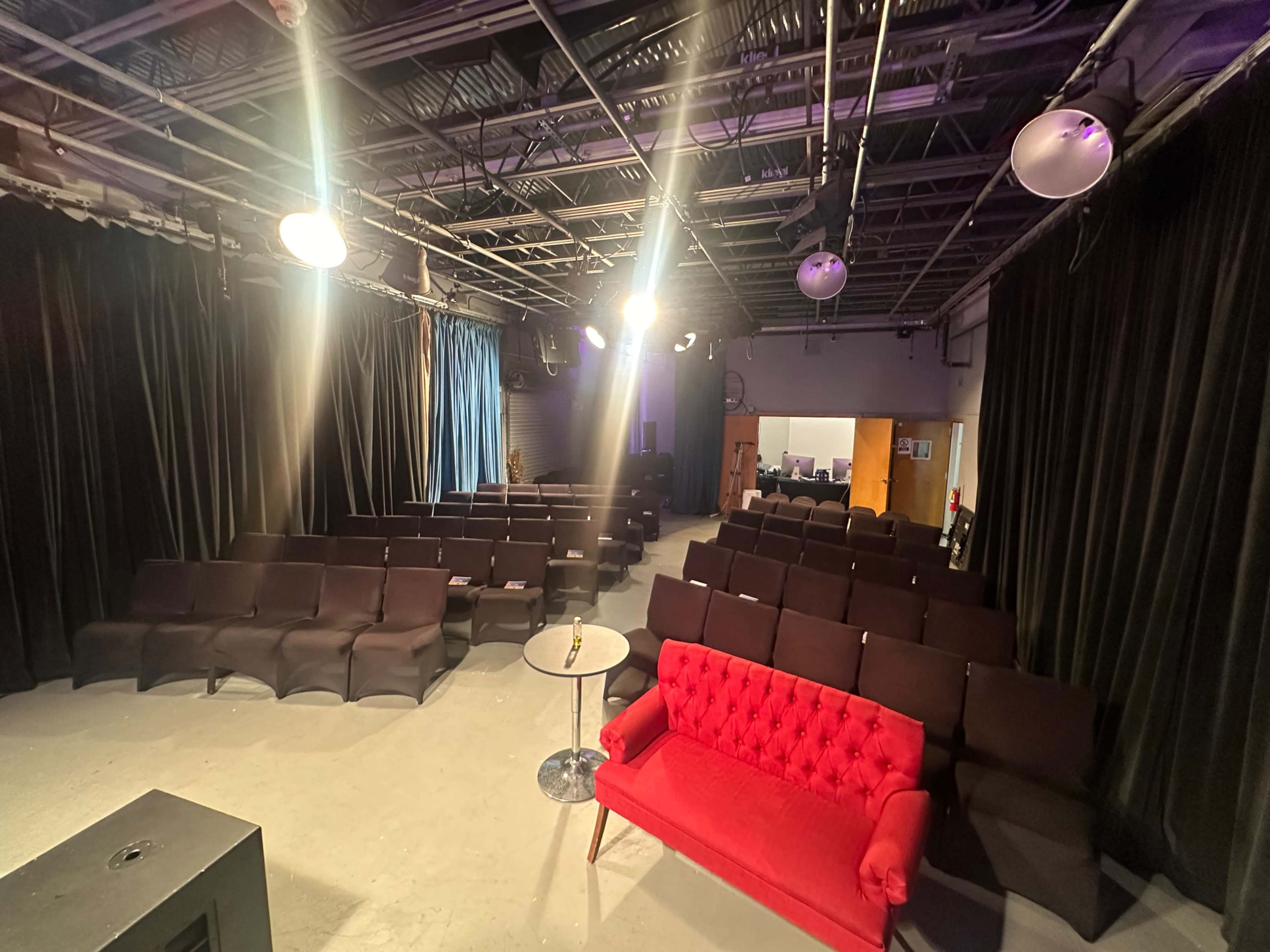 The image shows a small auditorium with rows of black chairs and a red couch in the foreground, illuminated by overhead lights.