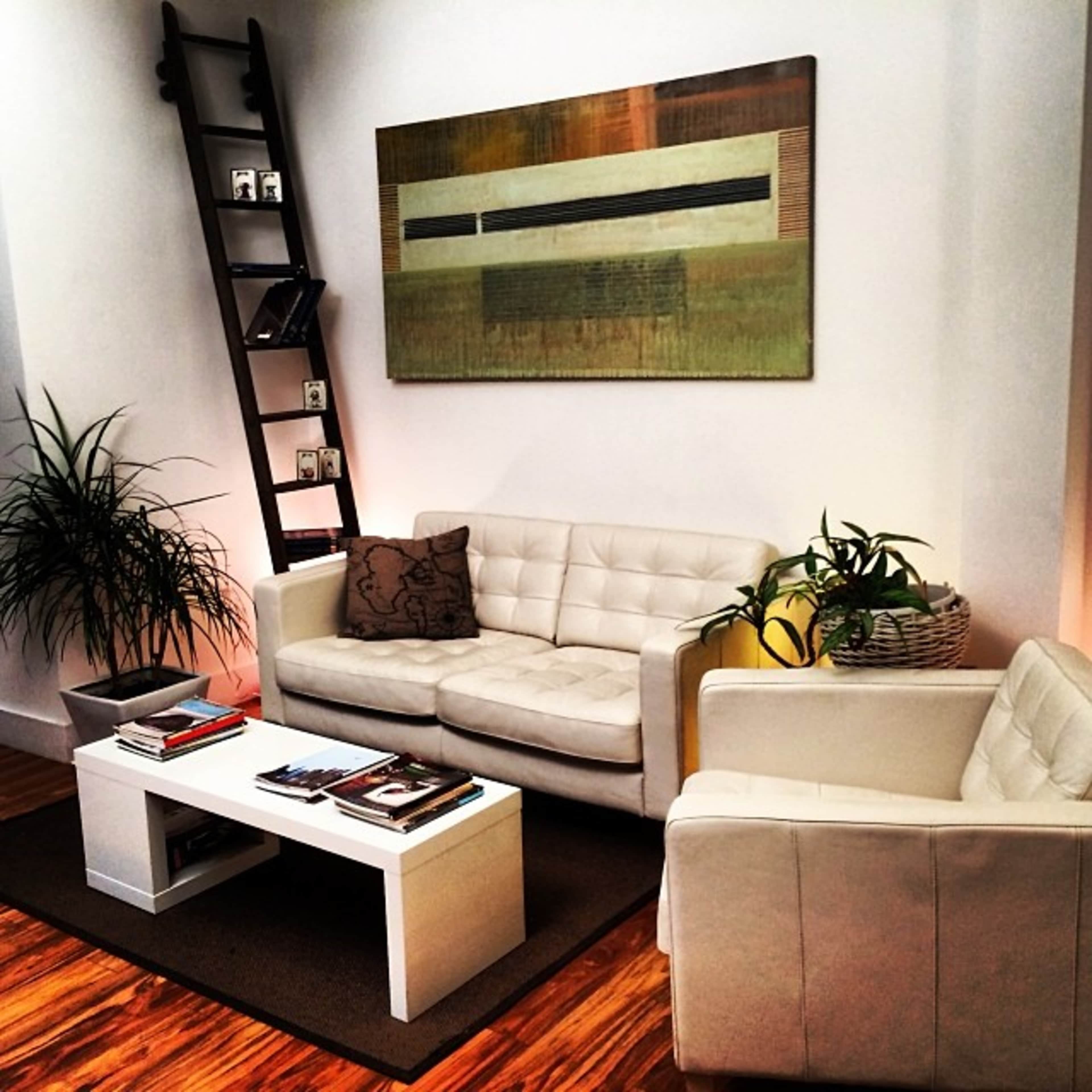 The scene features a modern living room with white leather sofas, a coffee table stacked with magazines, a ladder shelf holding framed photos, and potted plants.