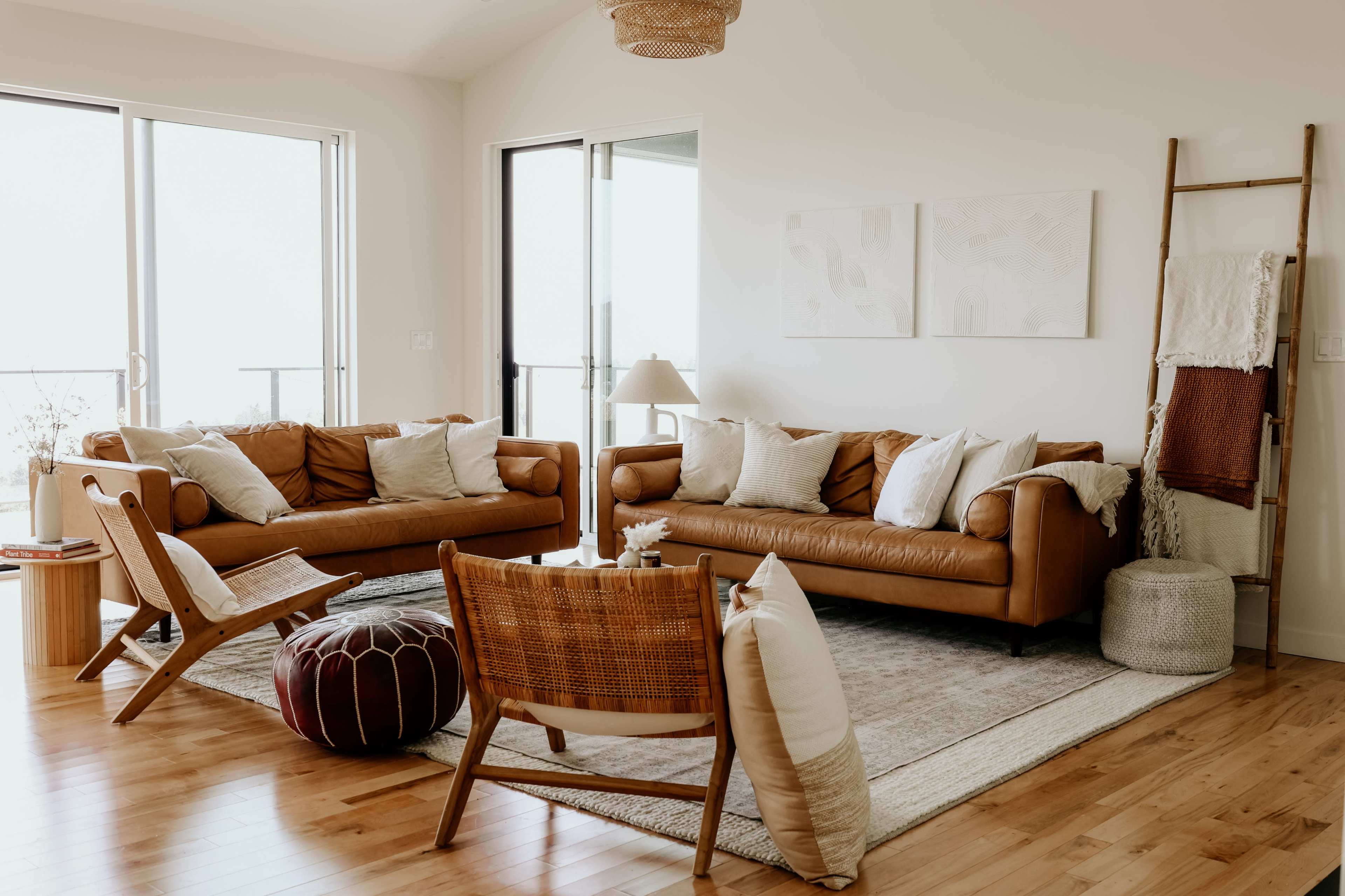 The living room features two brown leather sofas, a woven chair, a round pouf, and a wooden ladder displaying blankets, all arranged on a light-colored rug with large windows allowing natural light to enter.
