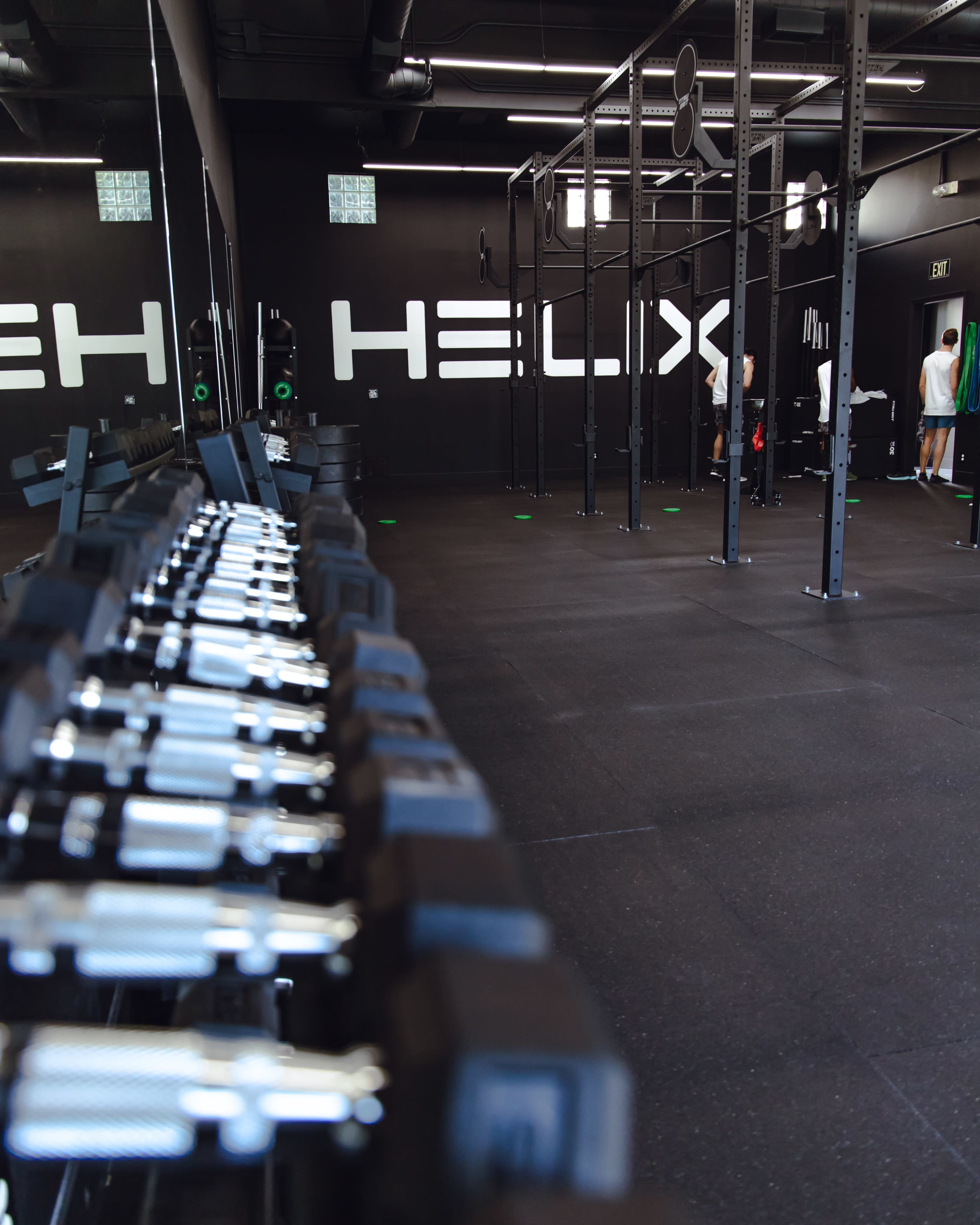 A gym interior with a row of dumbbells in the foreground and a functional training area in the background, featuring a large "HELIX" sign on the wall.