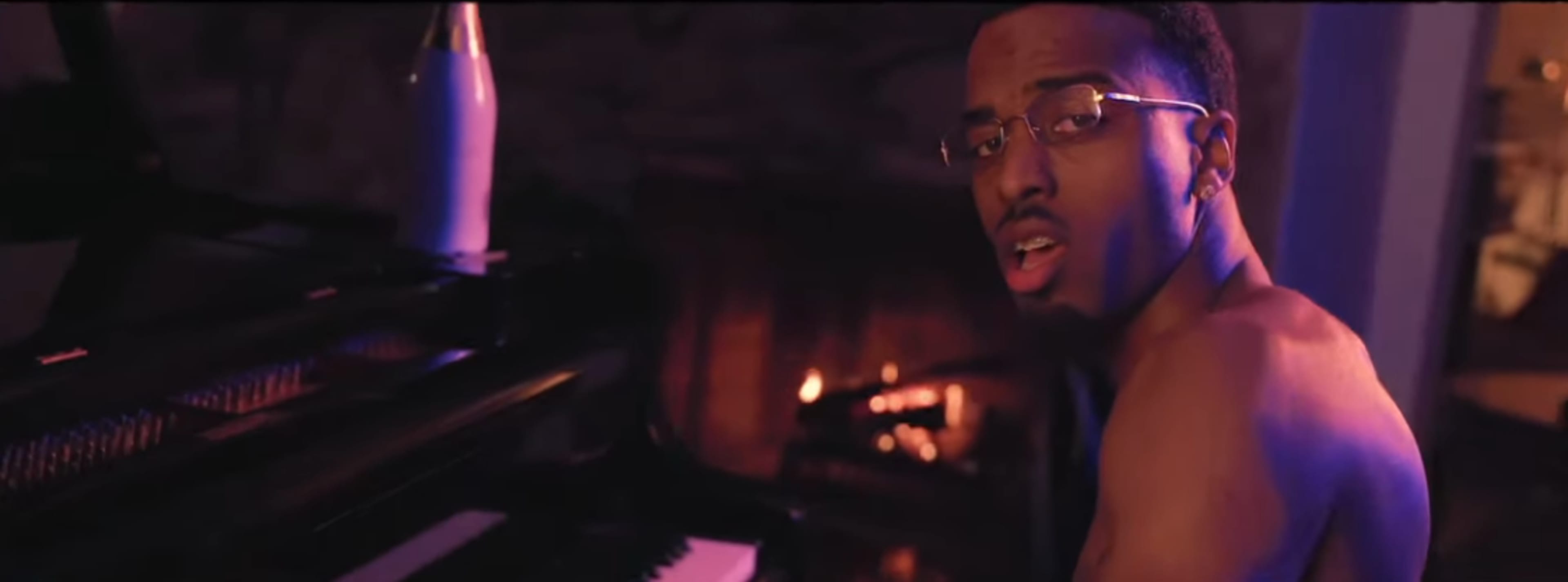 A shirtless man plays a piano in a dimly lit room with a fireplace.