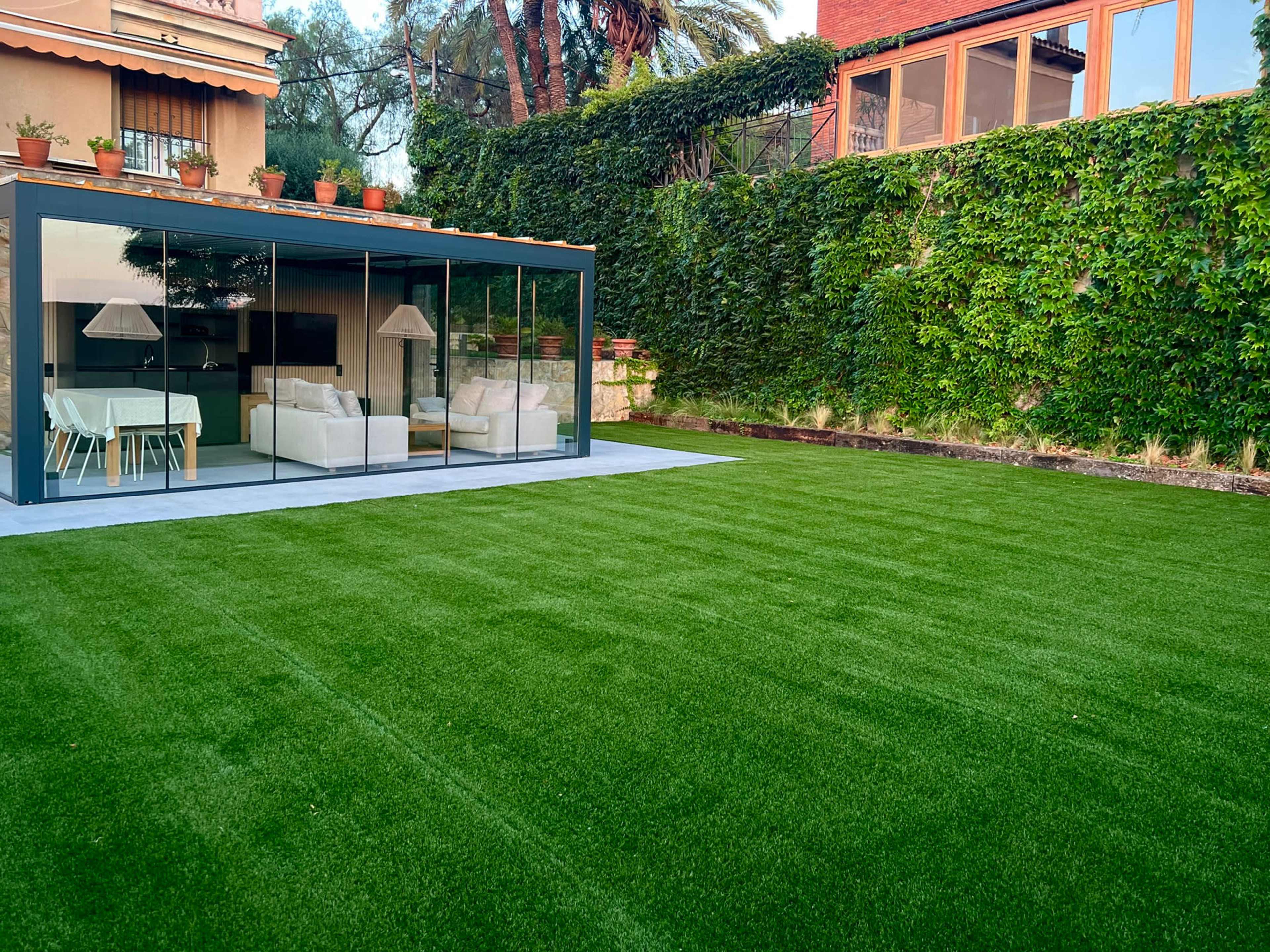 The image shows a modern glass-enclosed living space overlooking a green lawn with a wall of greenery in the background.