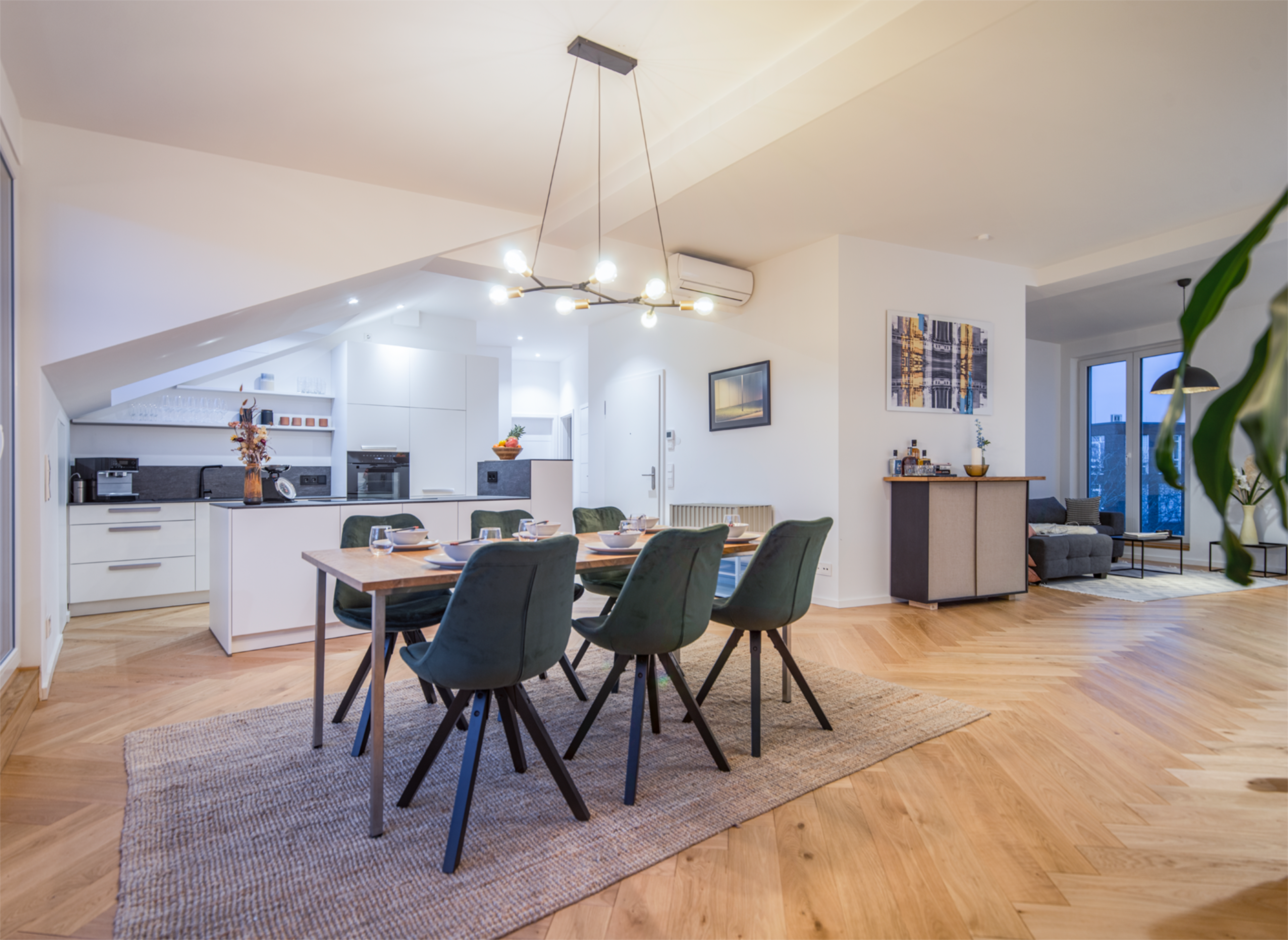 A modern kitchen and dining area featuring a long wooden table surrounded by green chairs, with sleek appliances in a well-lit space.