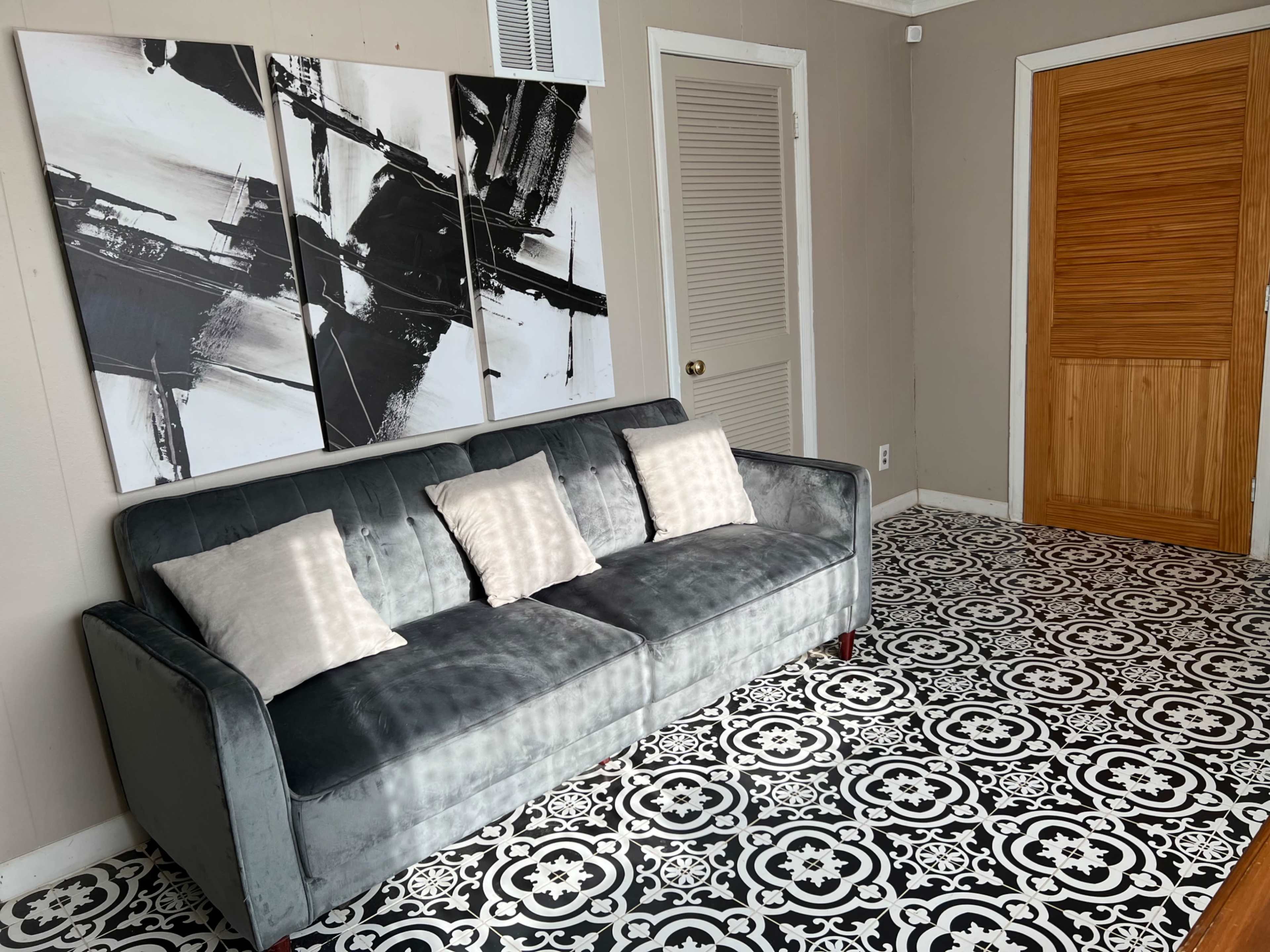 A living space featuring a gray sofa with two decorative pillows in front of a black and white patterned floor, accompanied by a series of abstract artwork on the wall and a wooden door.