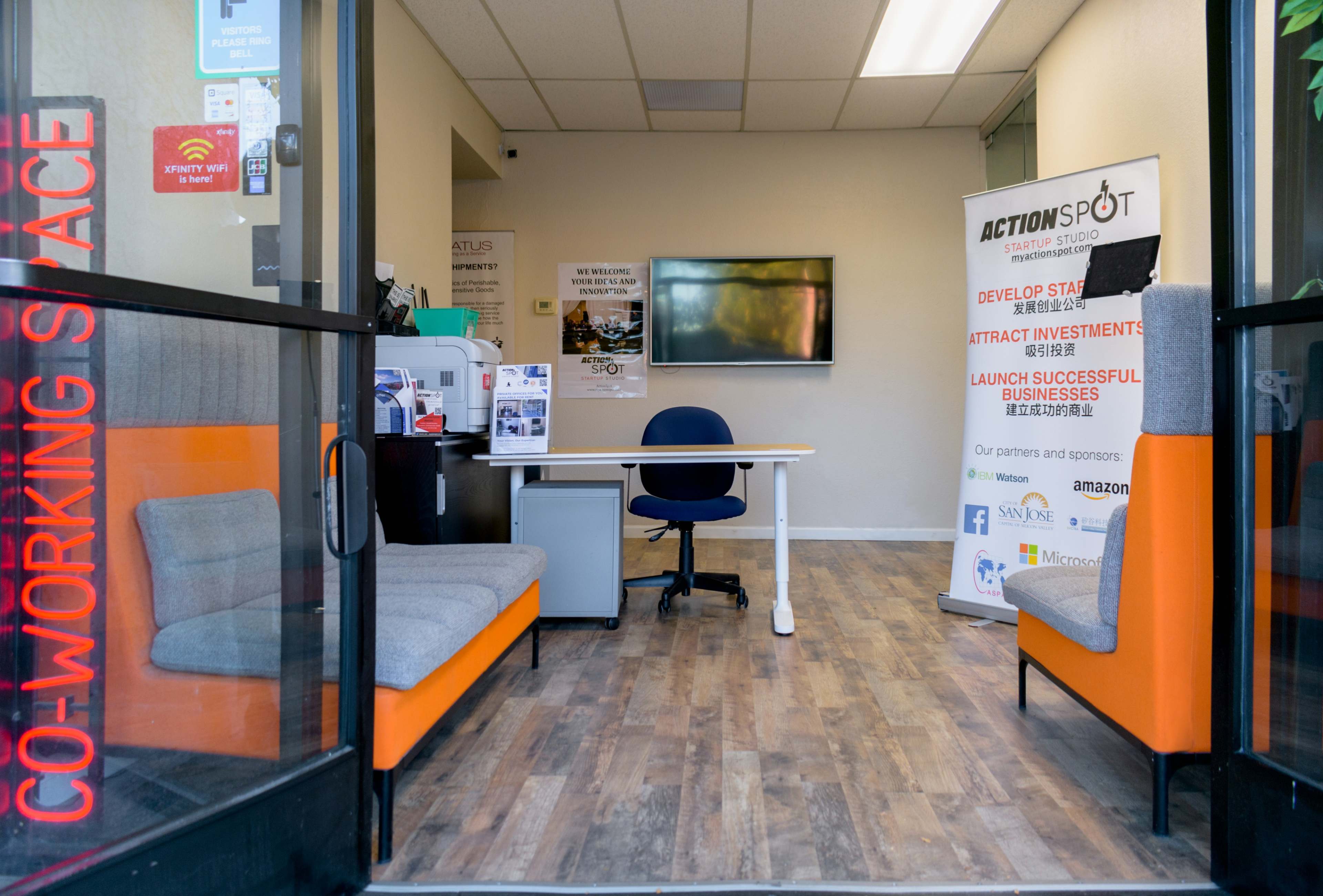 The image shows the entrance of a co-working space featuring a reception area with a desk, seating, and a promotional banner for ActionSpot.