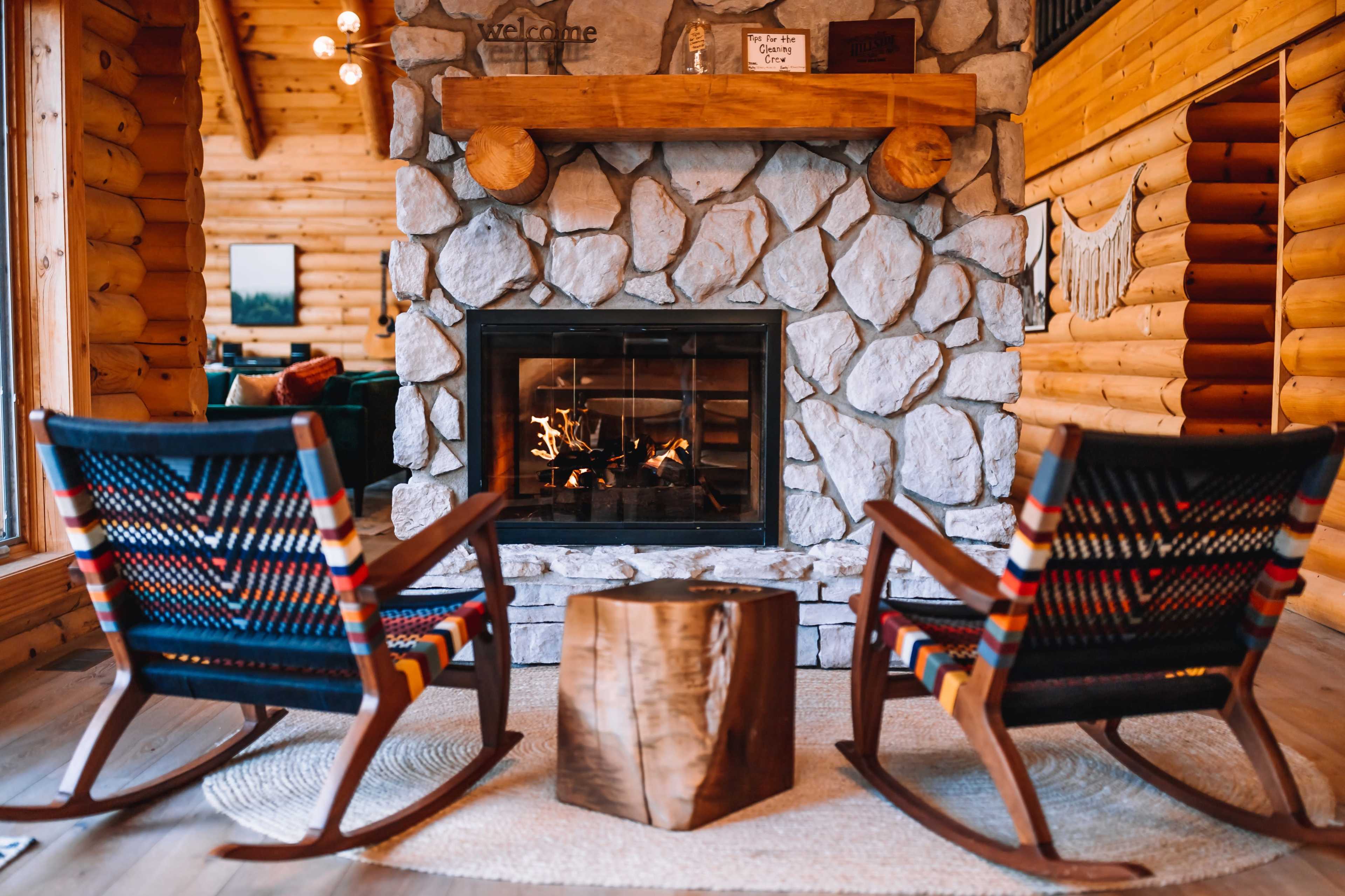 A cozy cabin interior featuring a stone fireplace flanked by two wooden rocking chairs and a small wooden table.