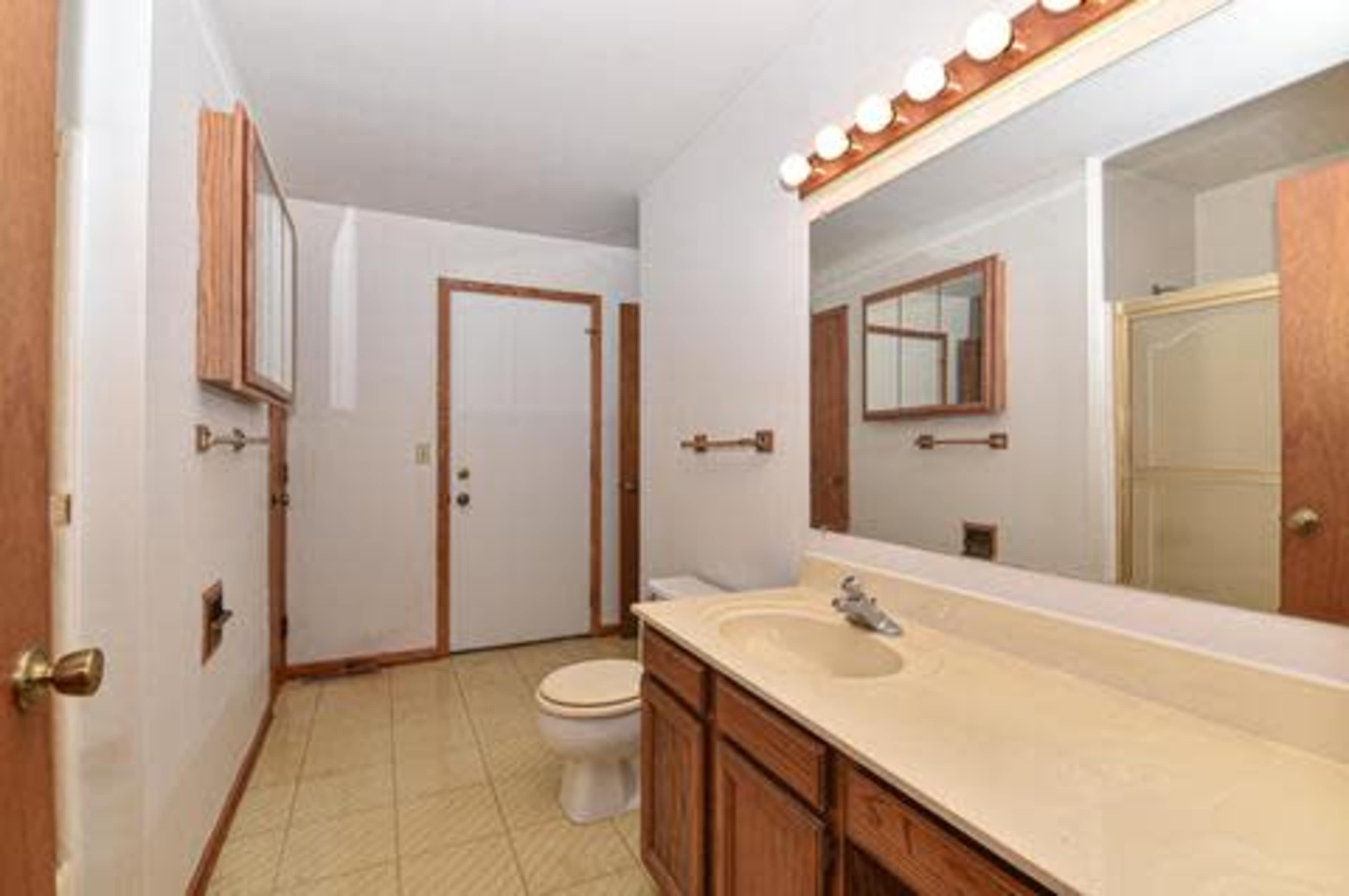 A simple bathroom featuring a toilet, a double sink vanity with wooden cabinets, a mirror above the sink, and a door leading outside.