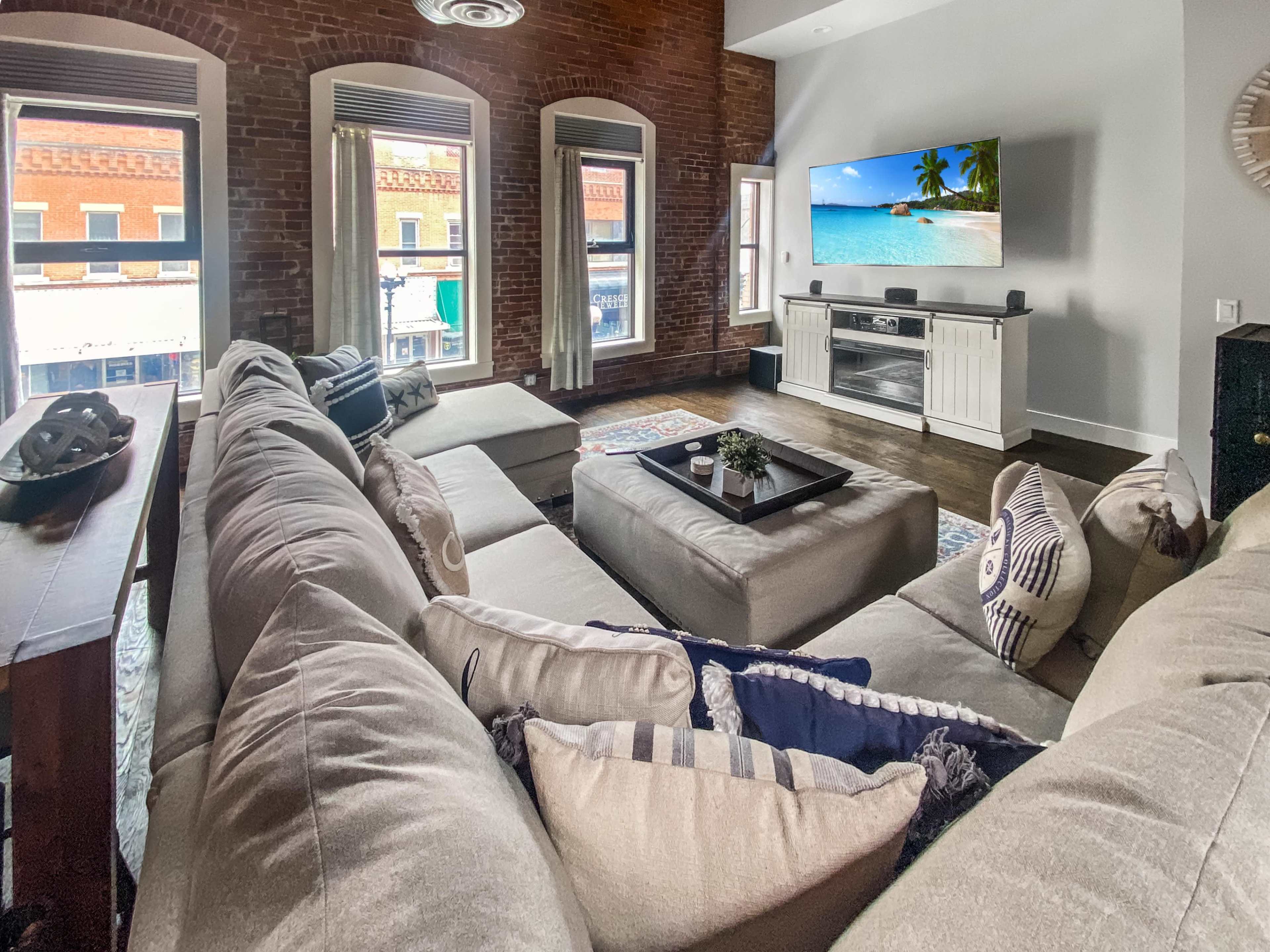 The living room features a large, U-shaped sofa, a coffee table in the center, and a television mounted on the wall, with exposed brick walls and large windows showing a view of the street outside.