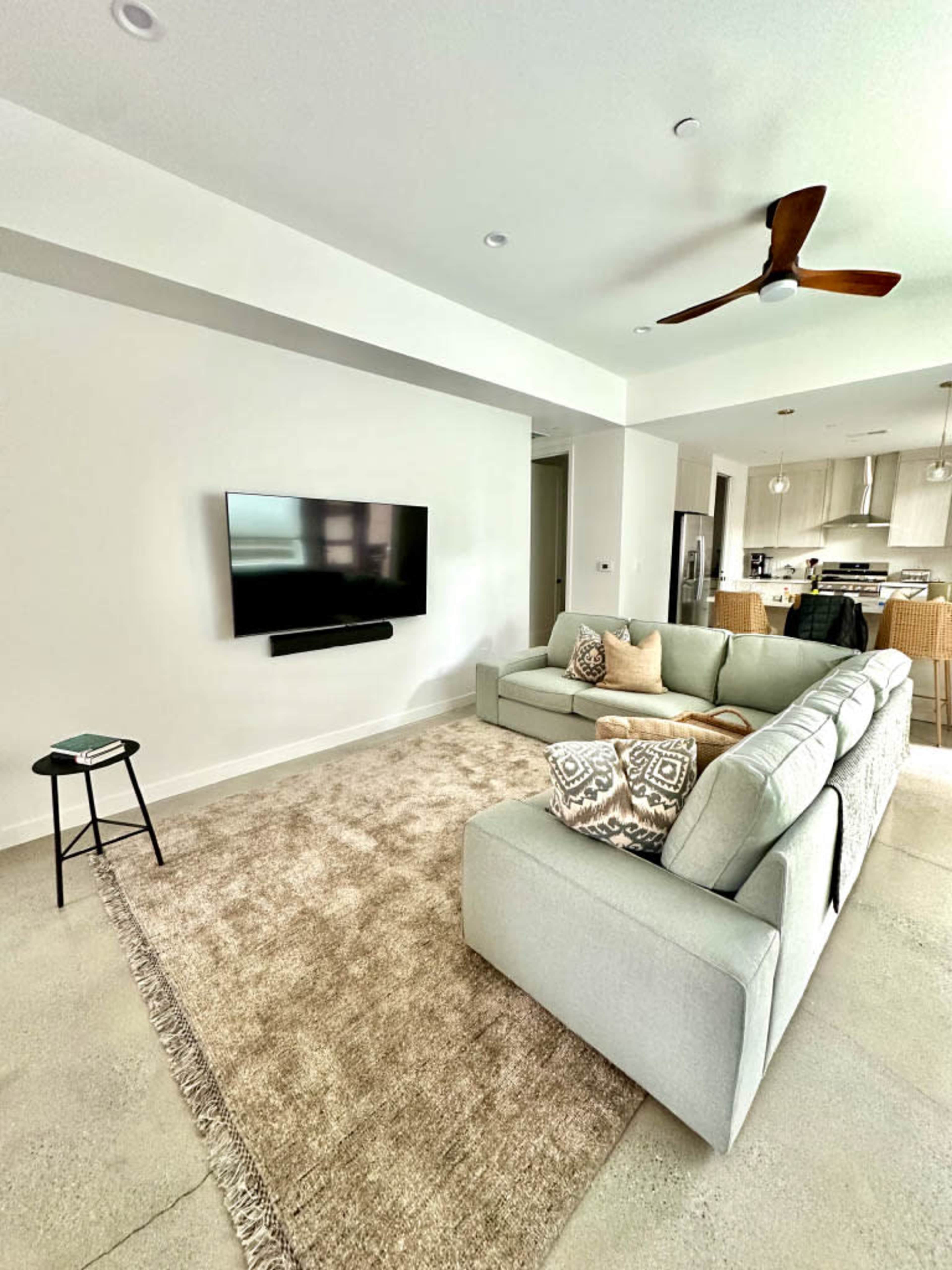 A light-colored living room features a sectional sofa, a wall-mounted television, and a carpet on a concrete floor, with a kitchen visible in the background.
