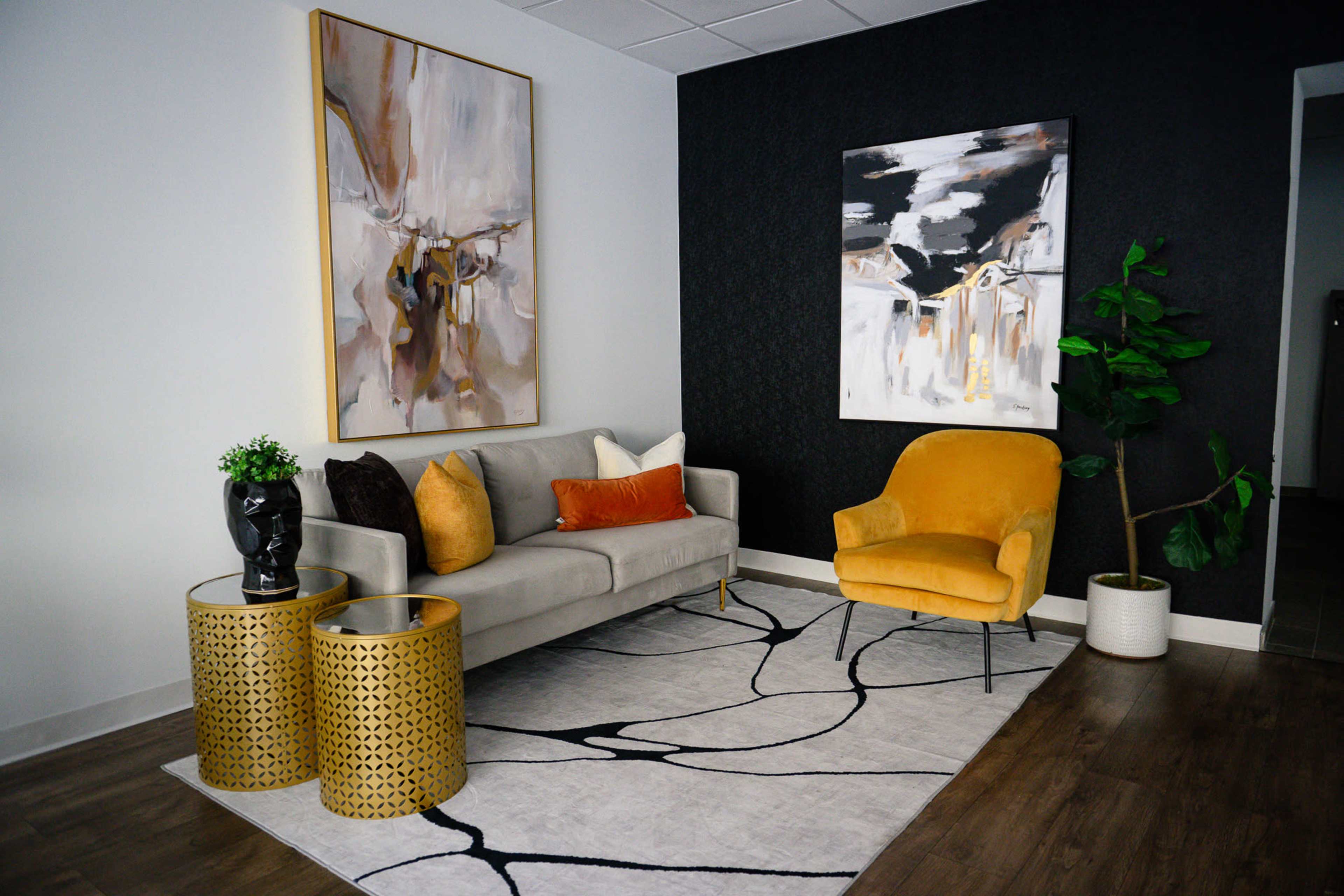 The image shows a modern seating area featuring a gray sofa with decorative pillows, a bright yellow chair, two gold patterned side tables, and two abstract paintings on the walls, all placed on a patterned rug.