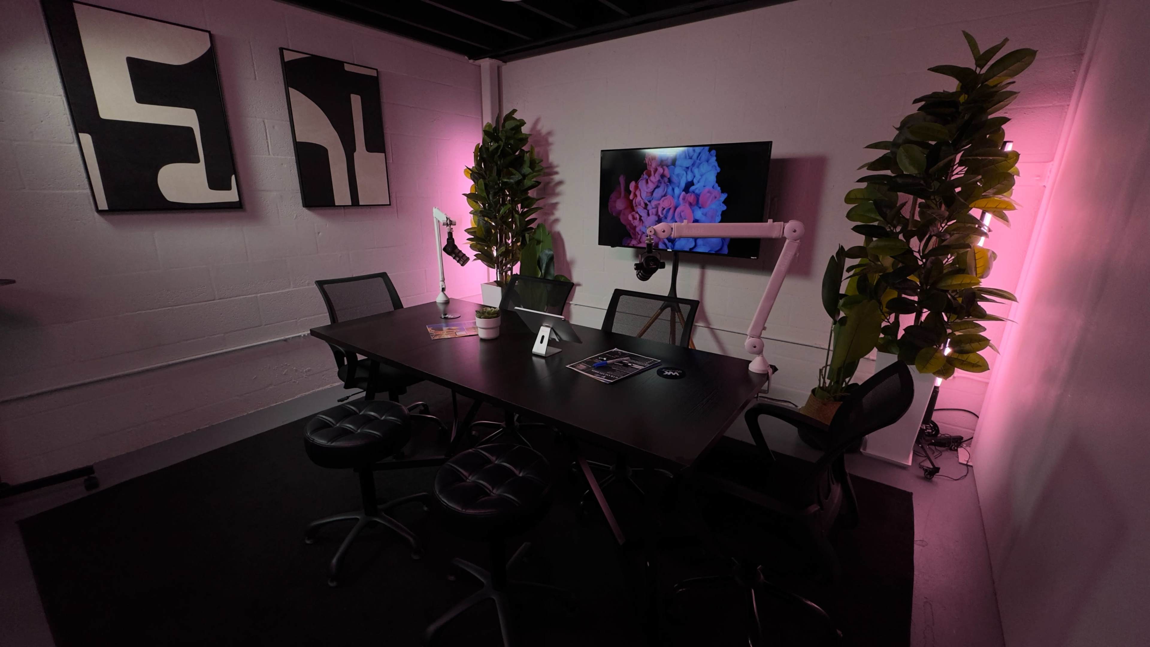 The image shows a modern meeting room with a black table surrounded by chairs, two large plants, a wall-mounted screen displaying colorful visuals, and abstract artwork on the walls, all illuminated by pink lighting.