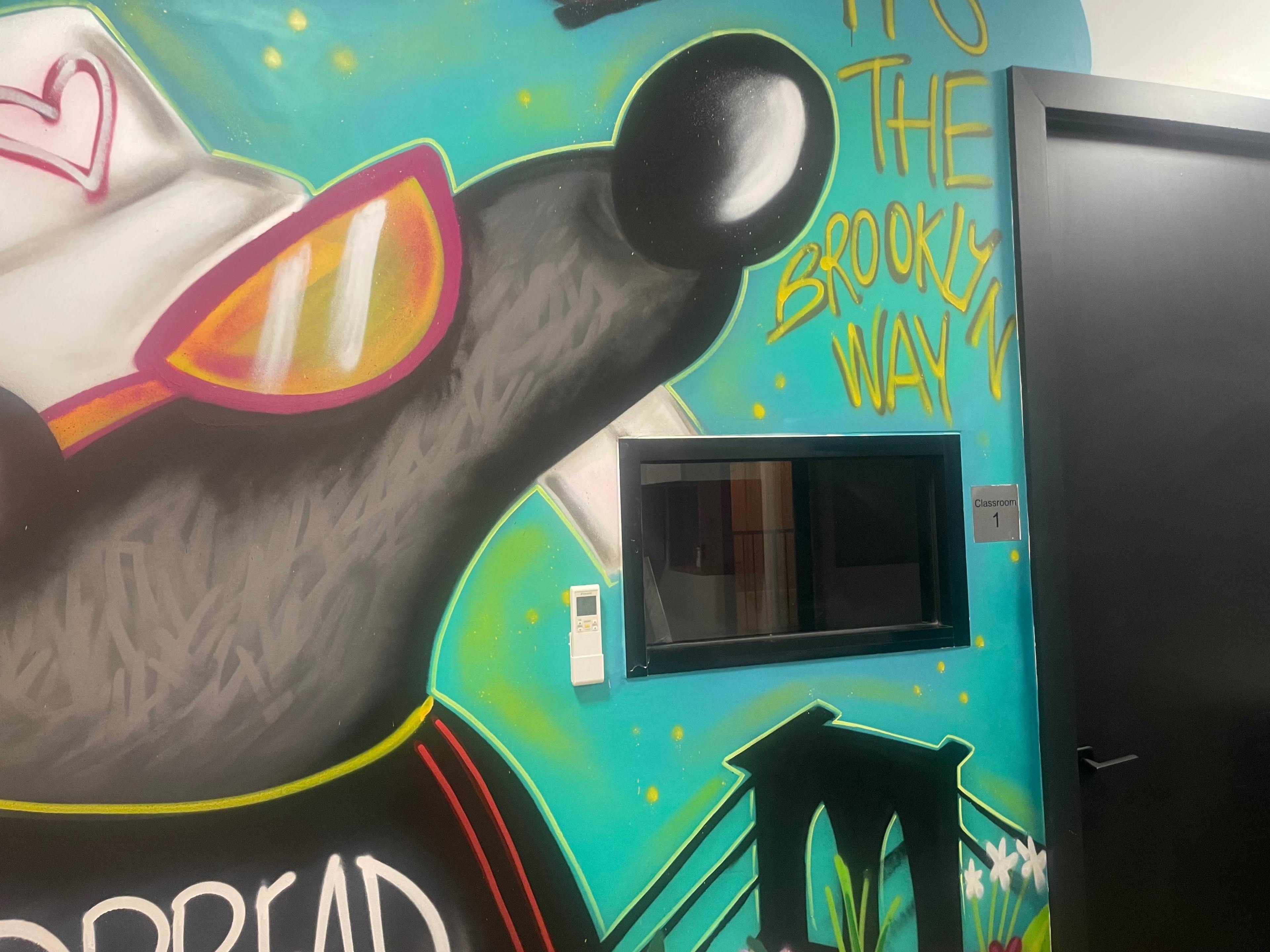 A colorful mural featuring a cartoon character wearing sunglasses and a cap is painted on a wall beside a window labeled "Classroom 1."