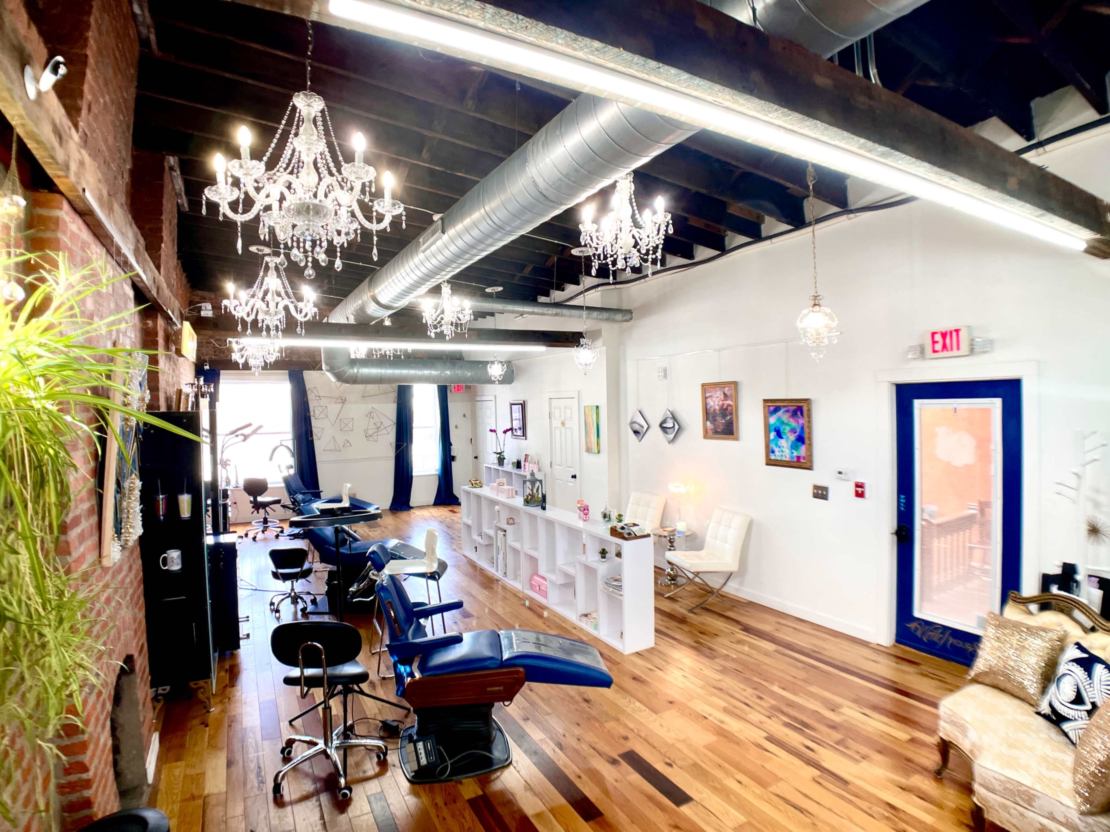 A spacious salon with exposed brick walls, wooden floors, and multiple styling stations under hanging chandeliers.