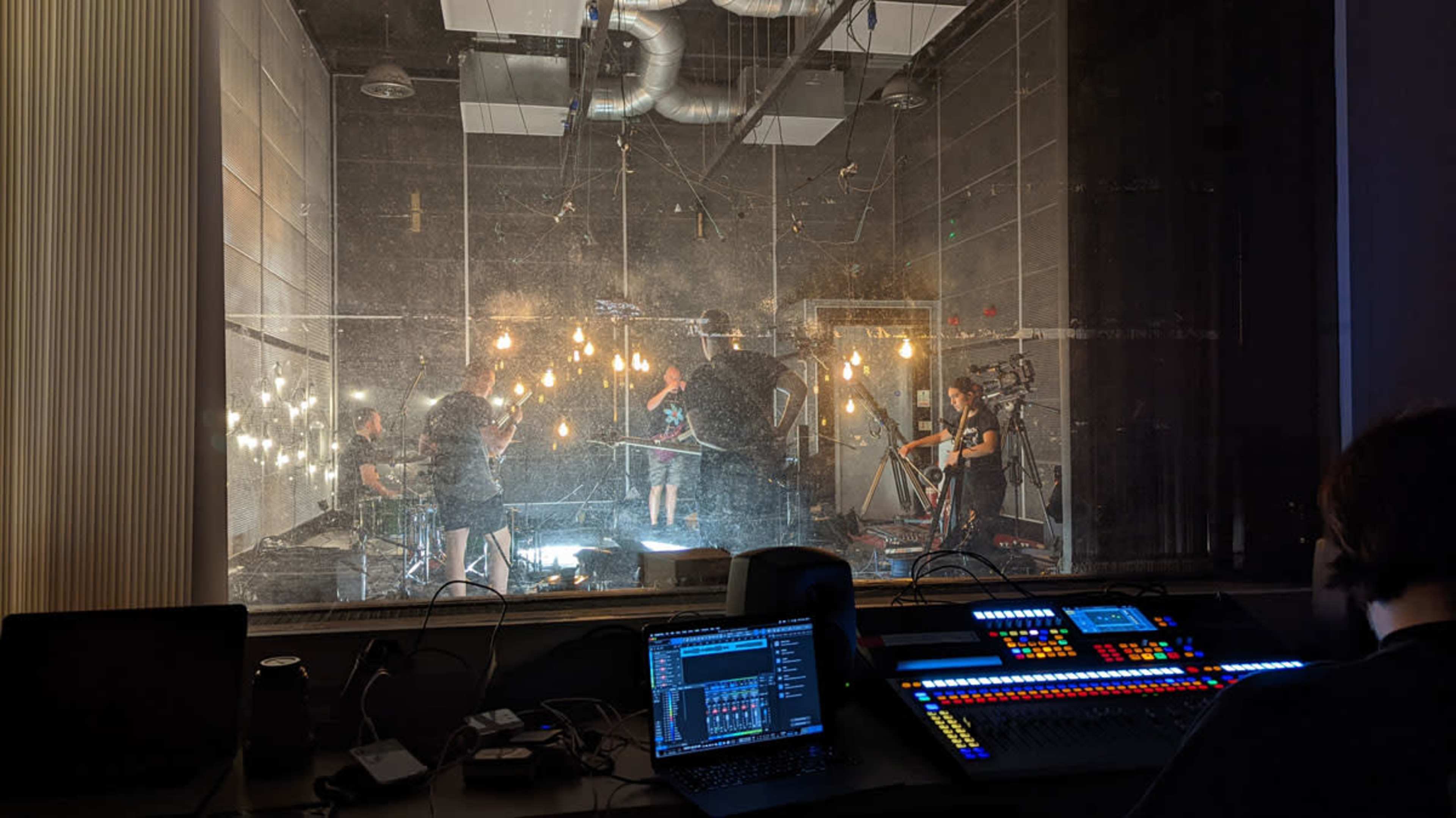 A control room overlooks a studio where several individuals are working with cameras and lighting equipment amid hanging bulbs.