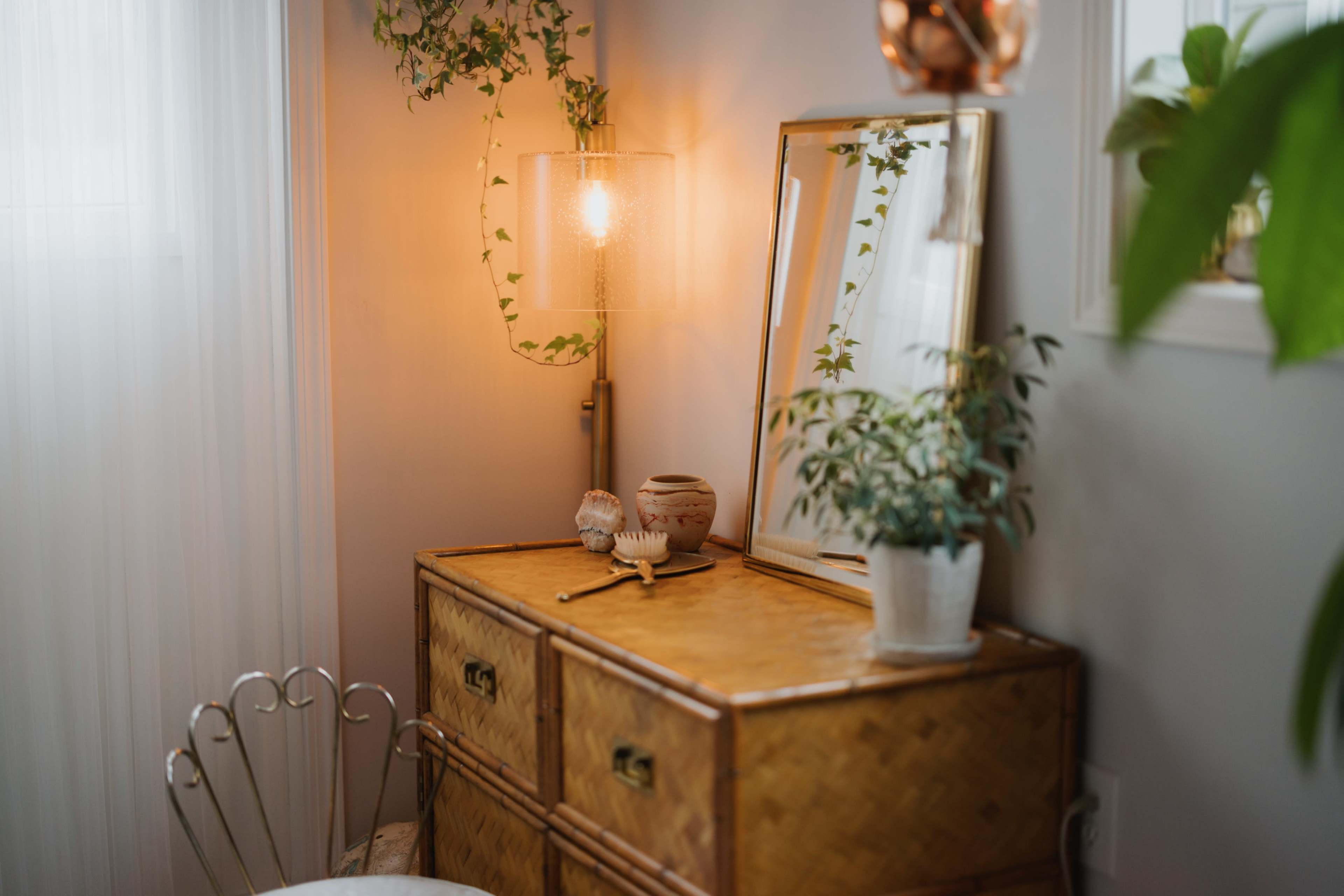 A woven storage chest with a mirror and decorative plants is positioned next to a window, illuminated by a warm light fixture.