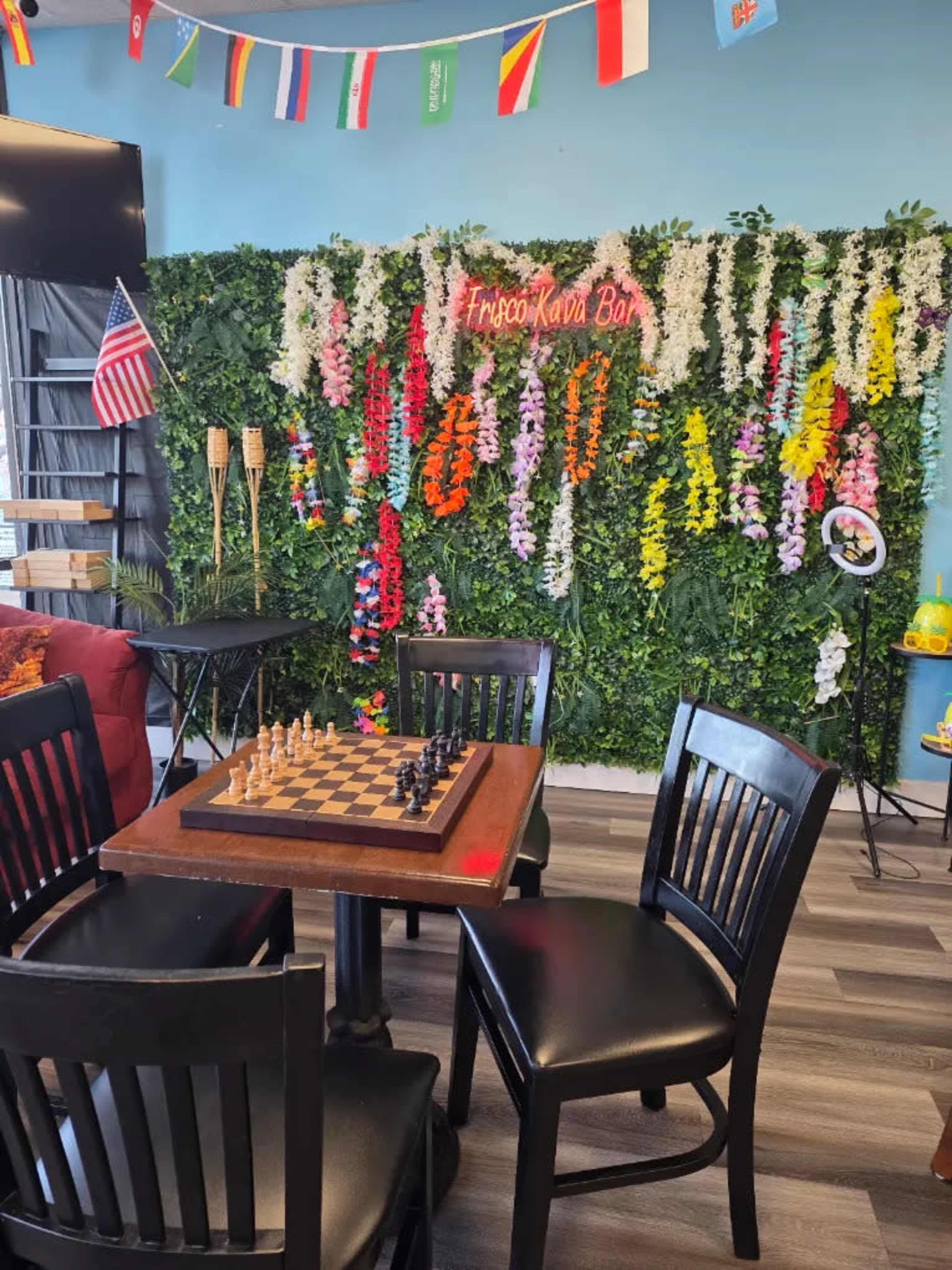 The image shows a cozy café setting with a chessboard on a wooden table, surrounded by chairs, and a vibrant floral wall adorned with colorful decorations and international flags.
