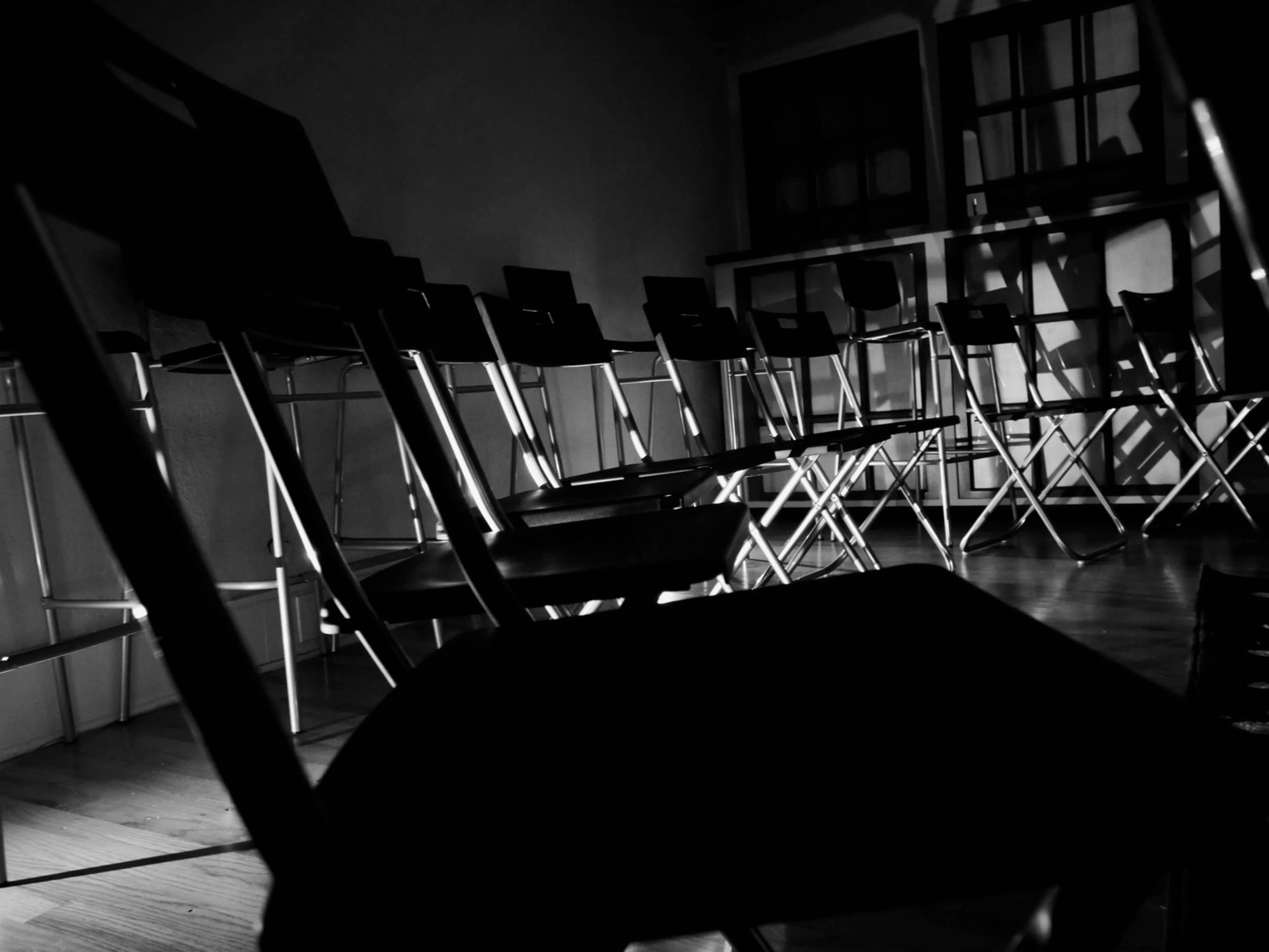 A circle of folding chairs casts shadows on a wooden floor in a dimly lit room.