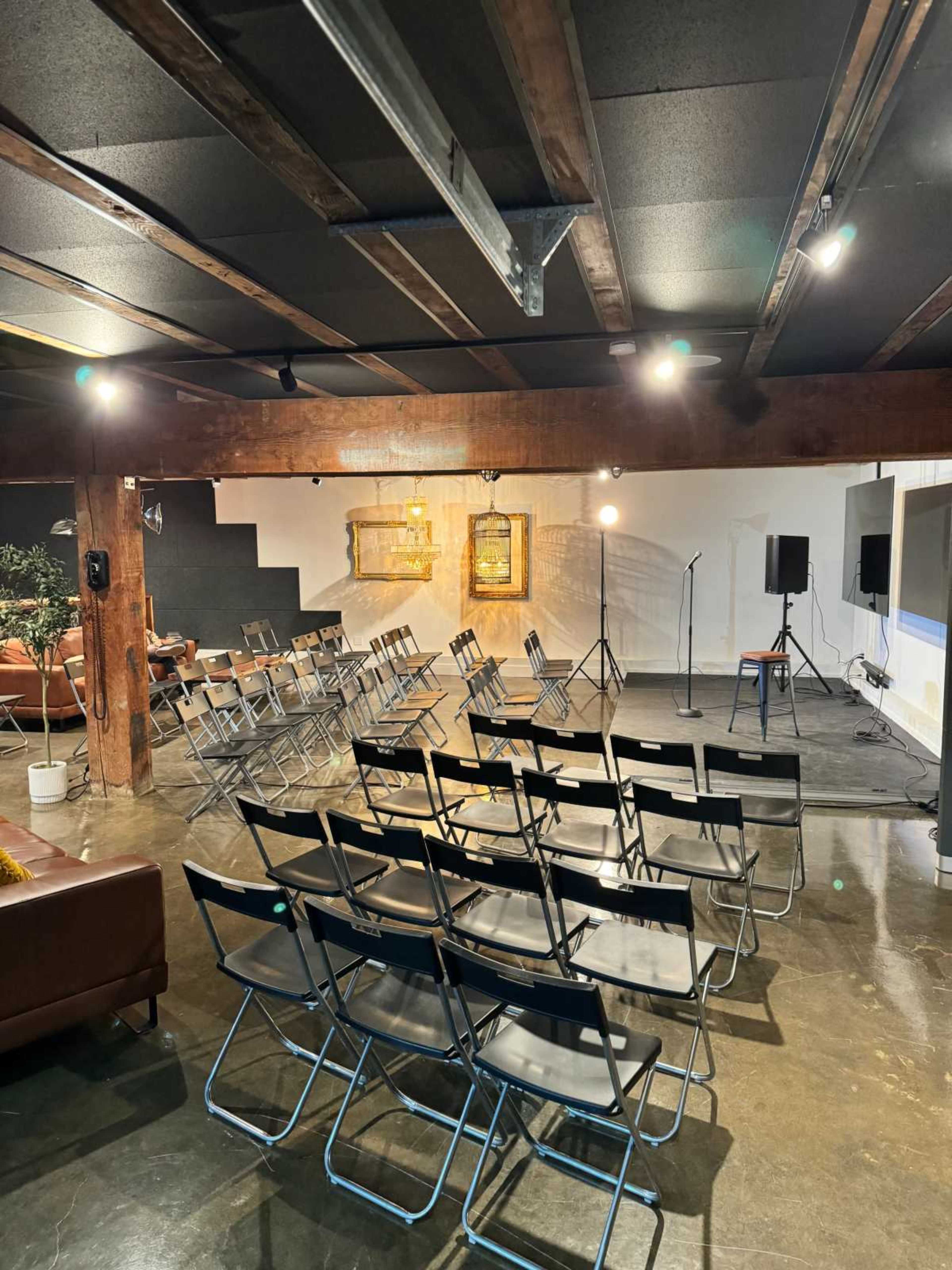 A spacious event room features rows of folding chairs facing a small stage with microphones and artwork on the walls.