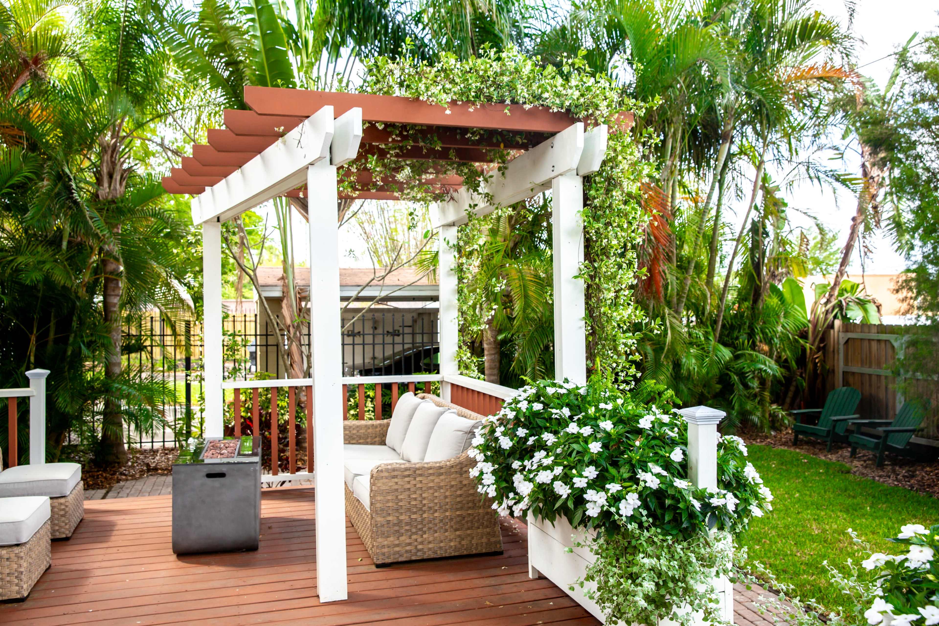A white pergola adorned with greenery and flowers provides shaded seating in a landscaped backyard surrounded by palm trees.