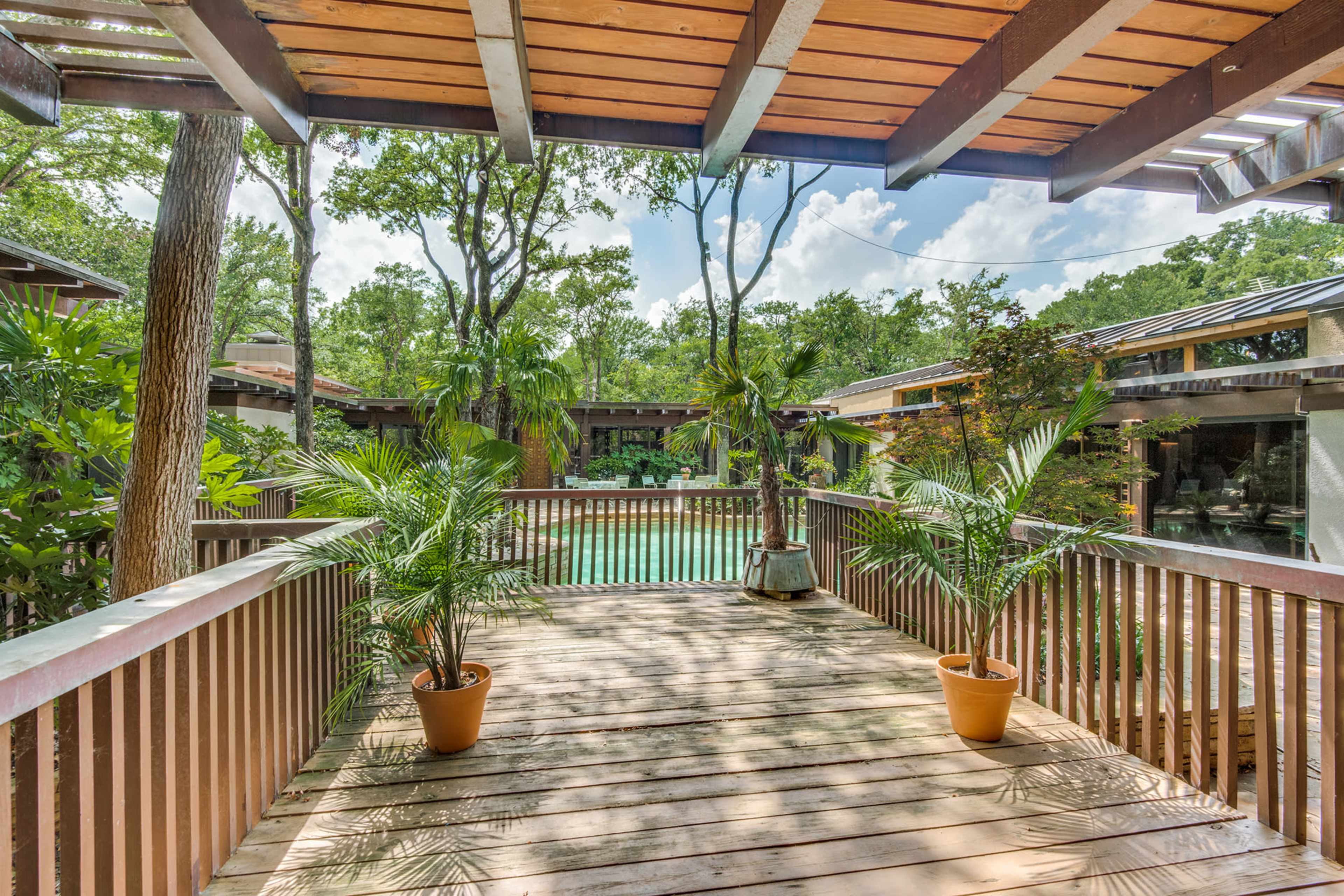 A wooden deck with potted plants overlooking a green pool area surrounded by trees and buildings.