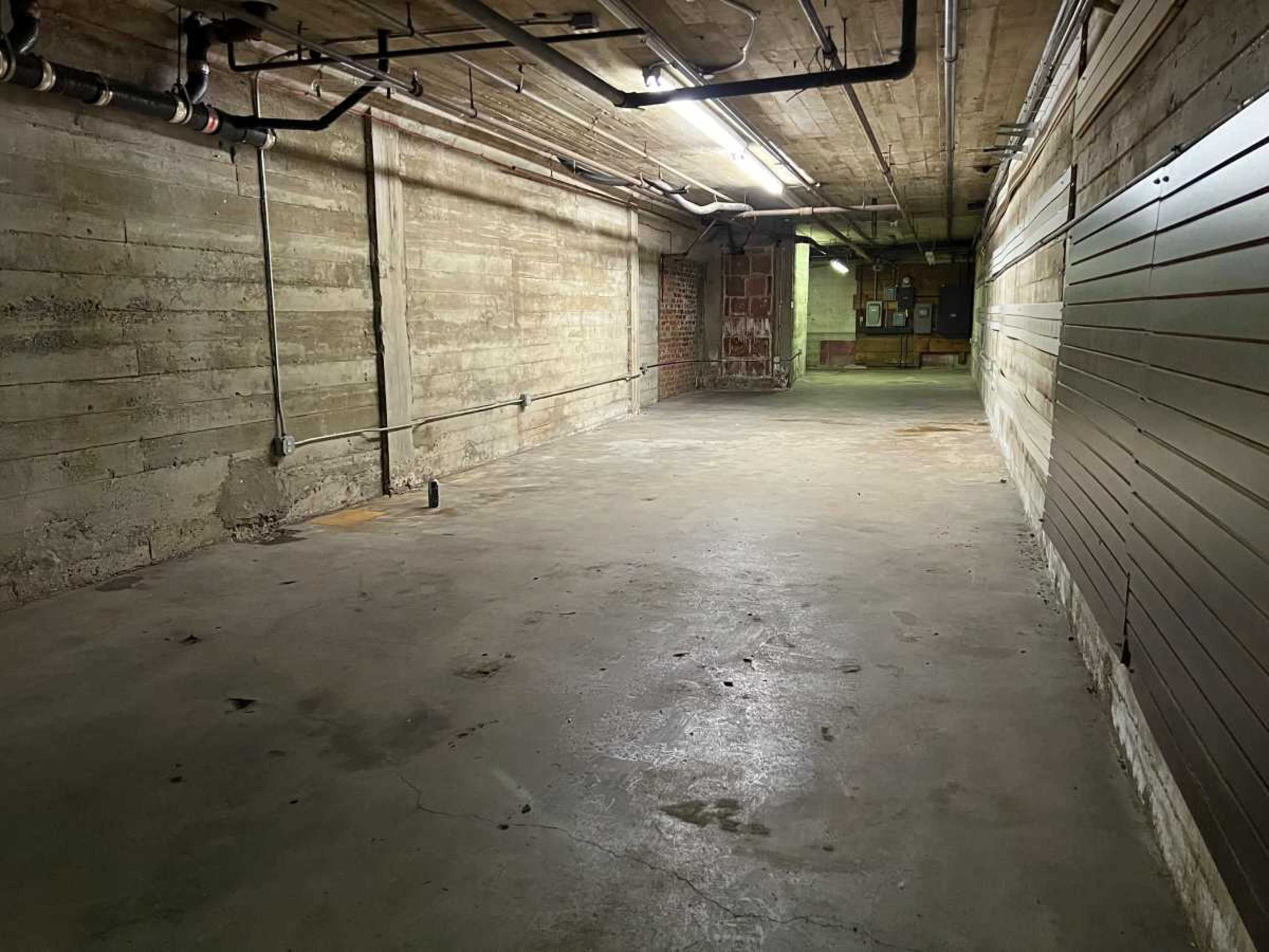 The image shows a wide concrete hallway with exposed pipes and brick walls, leading into a dimly lit area.
