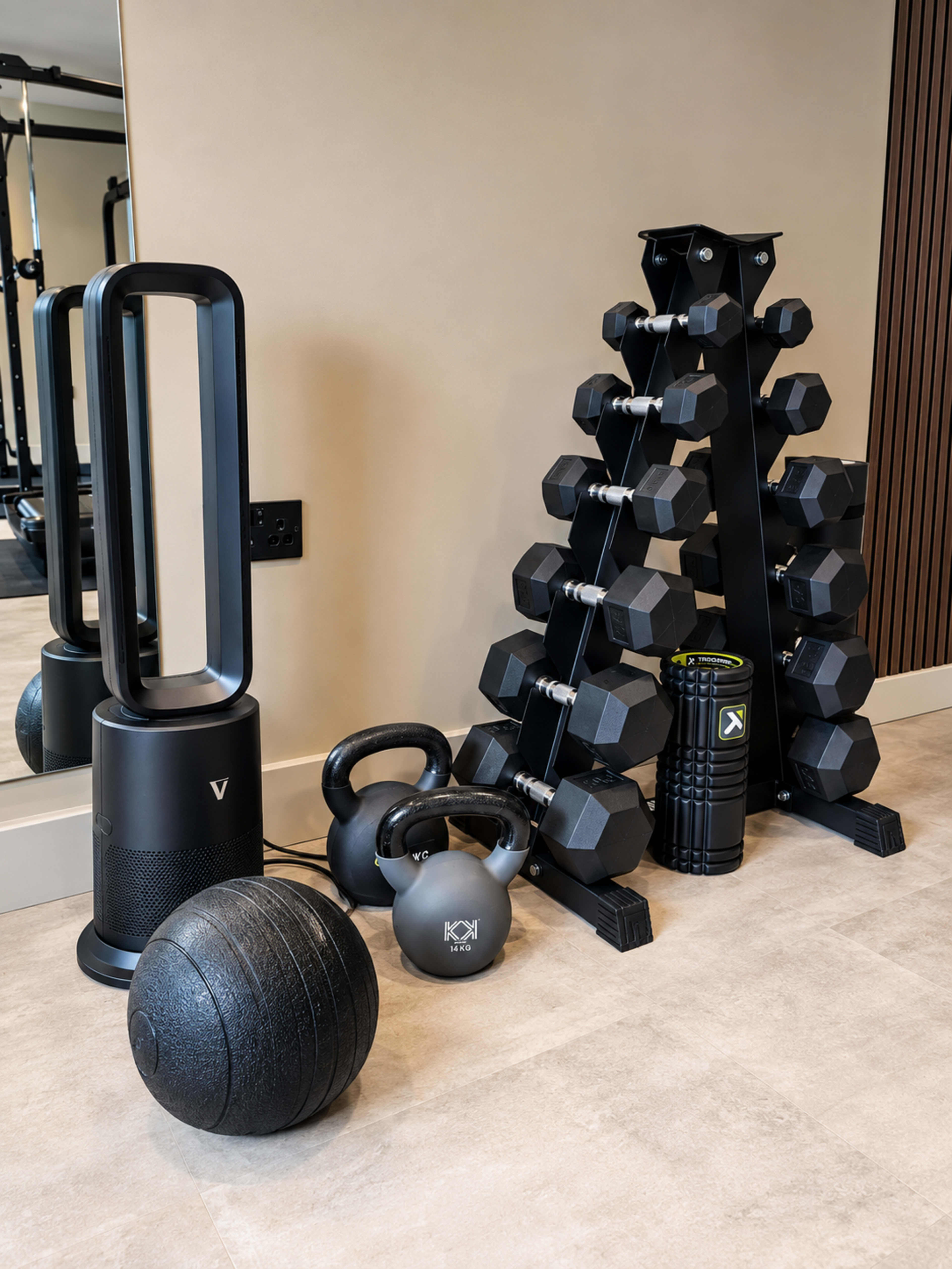 The image shows a home gym setup featuring a collection of weights, including dumbbells, kettlebells, a medicine ball, a foam roller, and a fan.