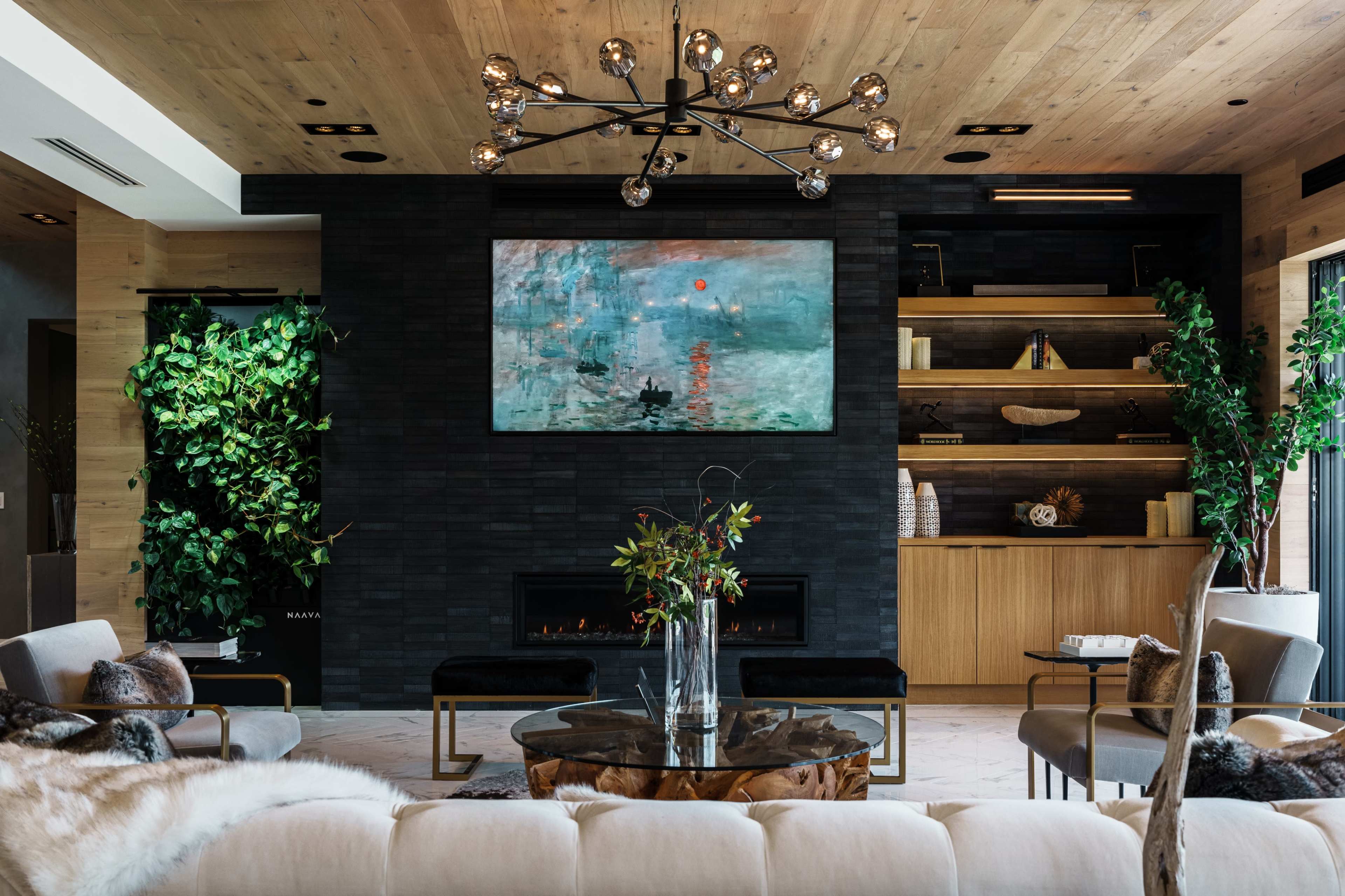 A modern living room features a large abstract painting above a sleek black fireplace, surrounded by greenery and stylish furniture.