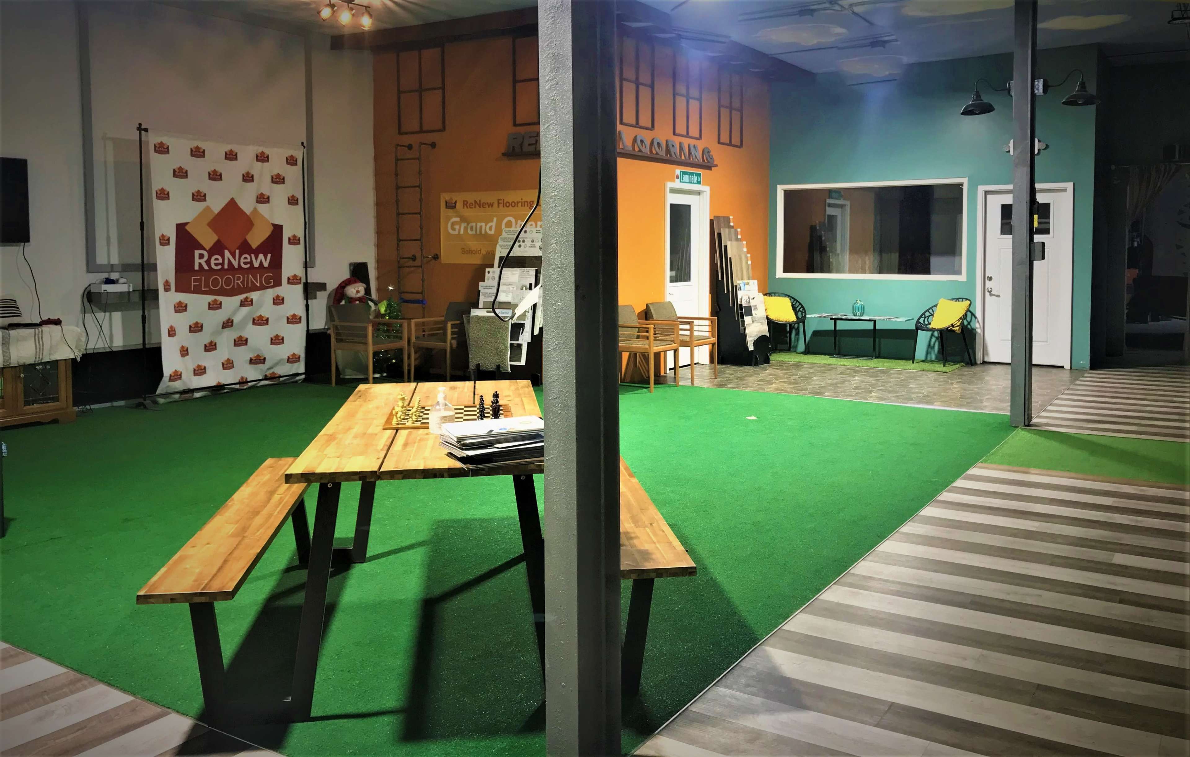 A spacious indoor area with green artificial grass, featuring a long picnic table with a chess set, surrounded by colorful walls and seating arrangements.