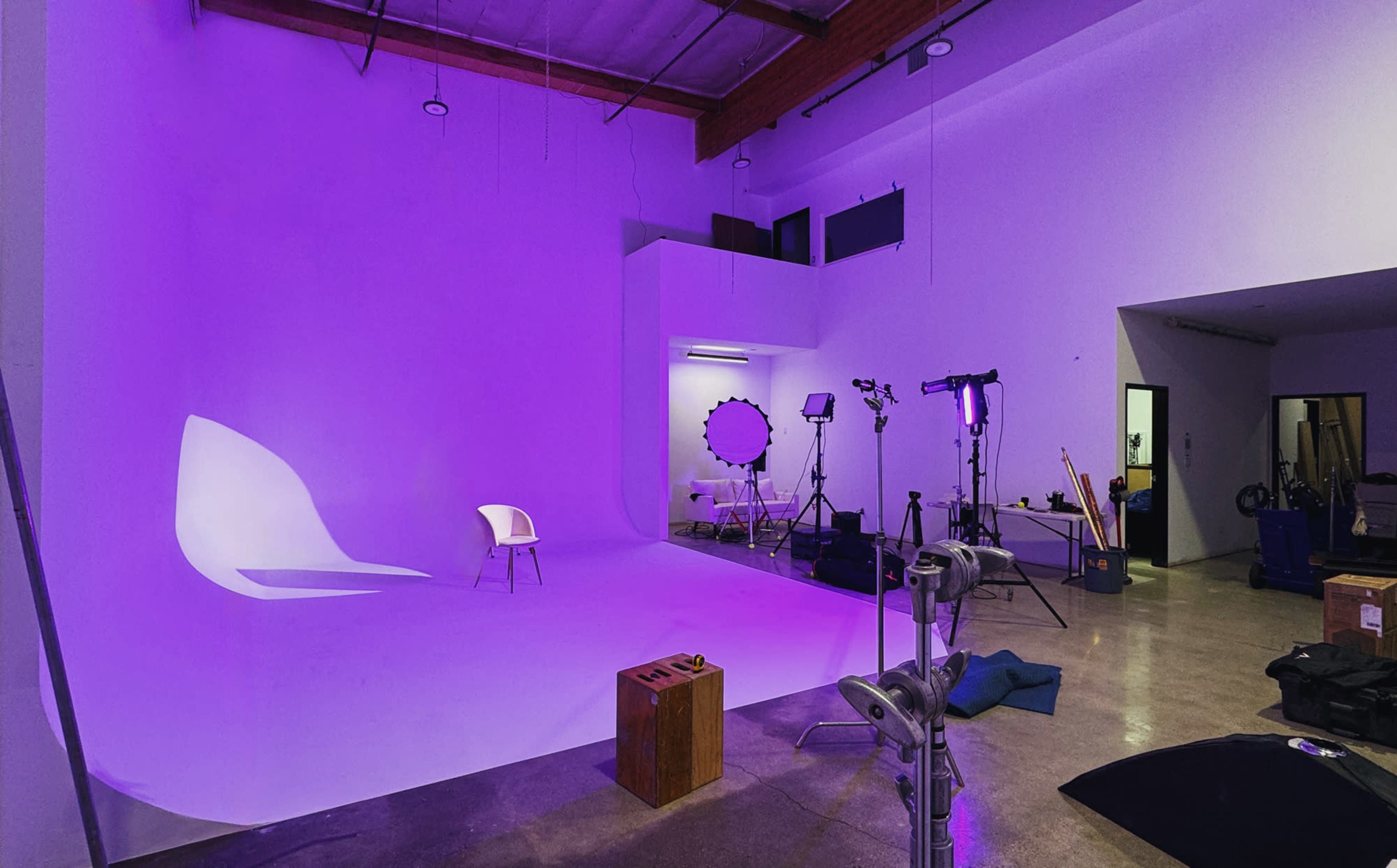 A photography studio features a purple backdrop, a white chair, various lighting equipment, and scattered props.