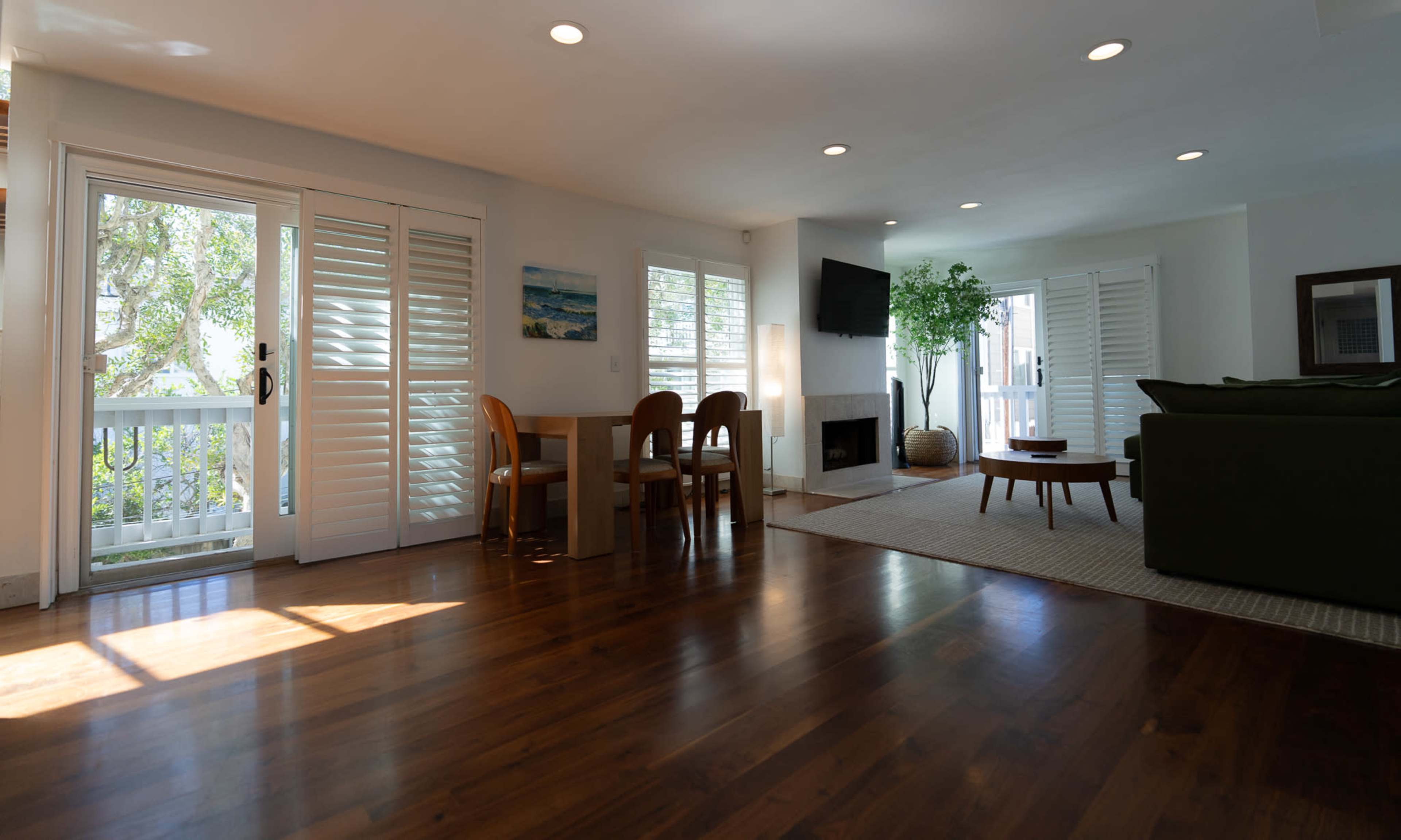 The image shows a bright and open living space with wooden flooring, a dining area, a green sofa, and large windows letting in natural light.