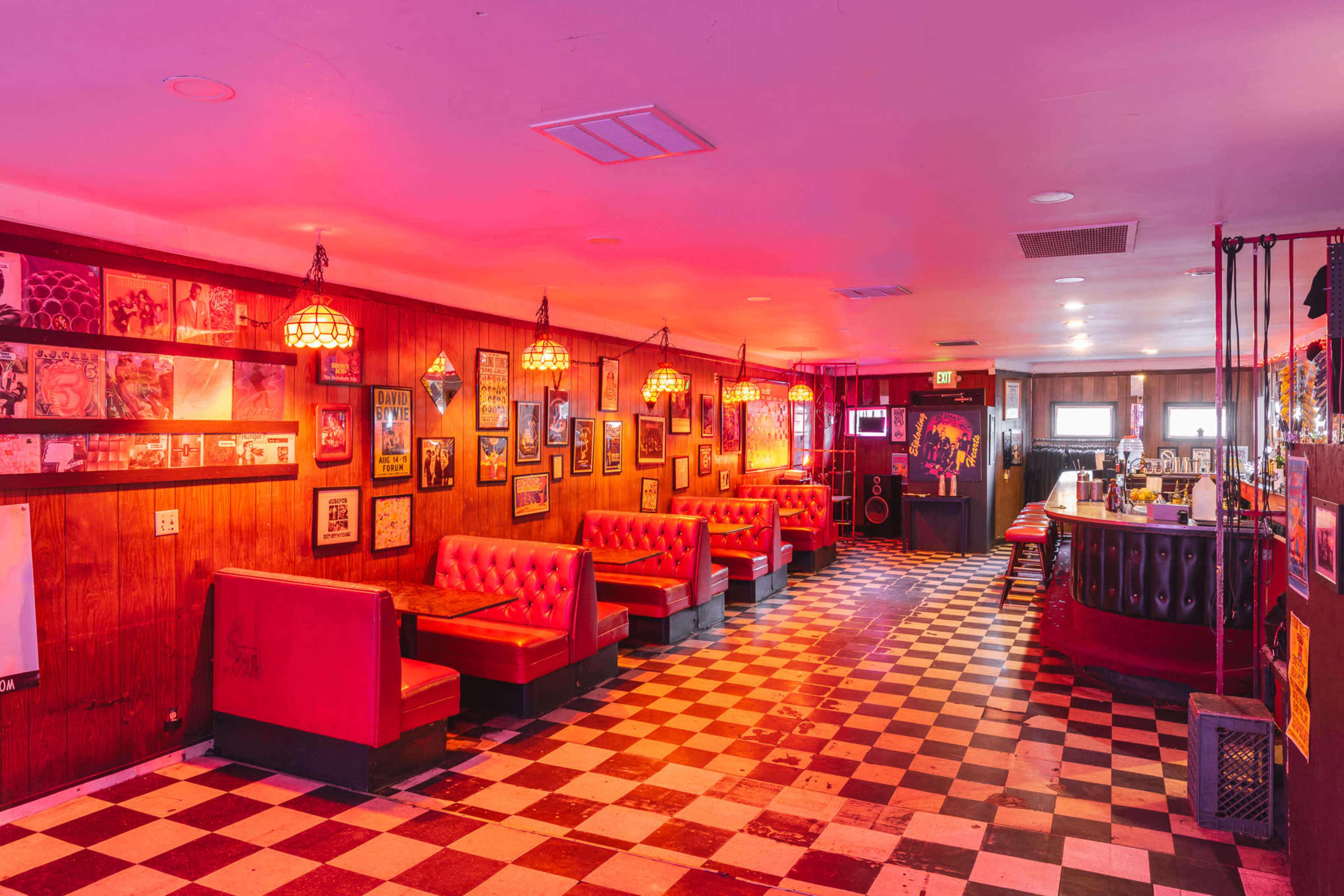 A retro-style diner interior featuring red booths and a black-and-white checkered floor, illuminated by warm lighting and adorned with framed artwork on the walls.