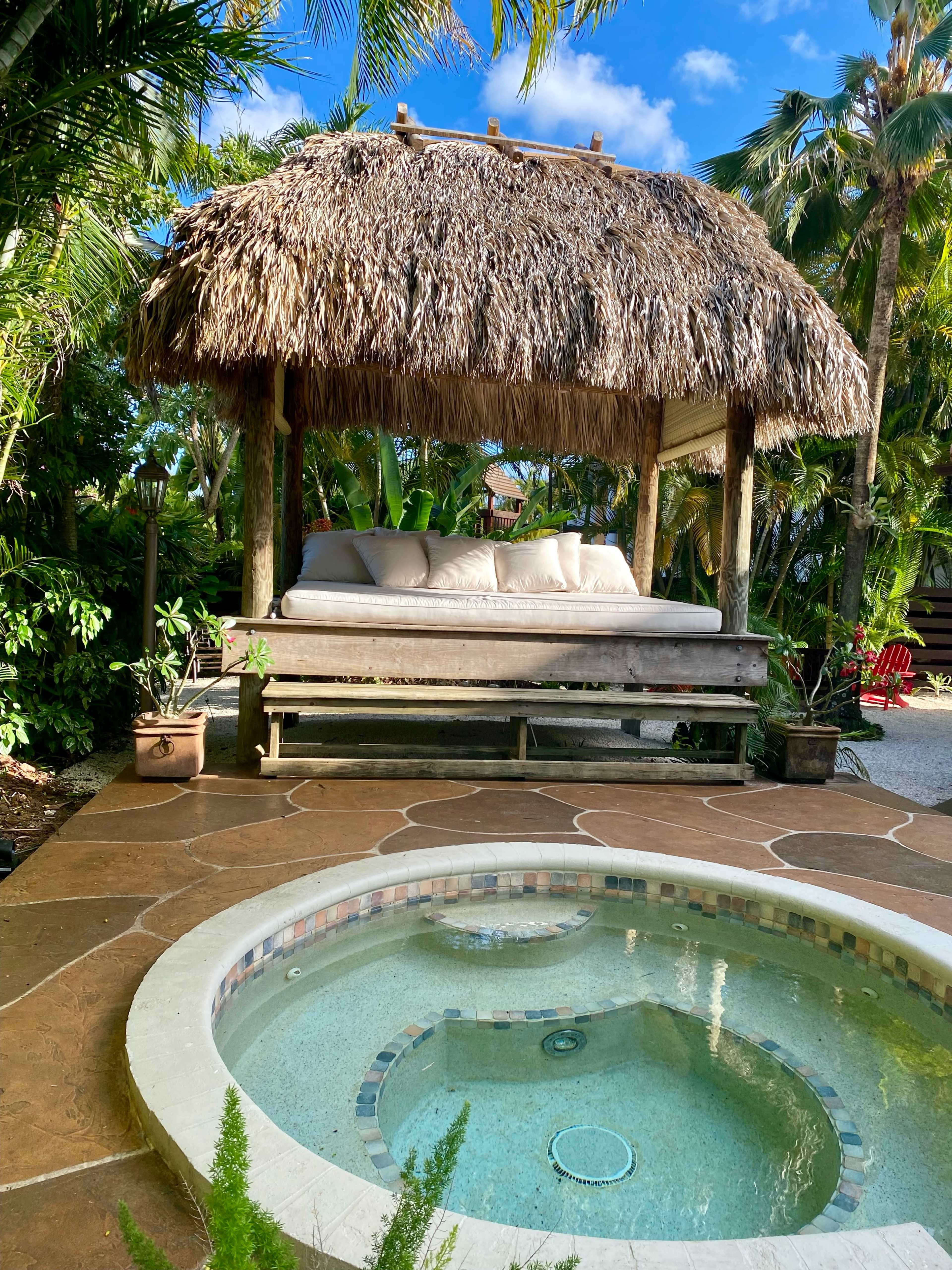 A thatched gazebo with a cushioned bench overlooks a circular hot tub surrounded by tropical plants.